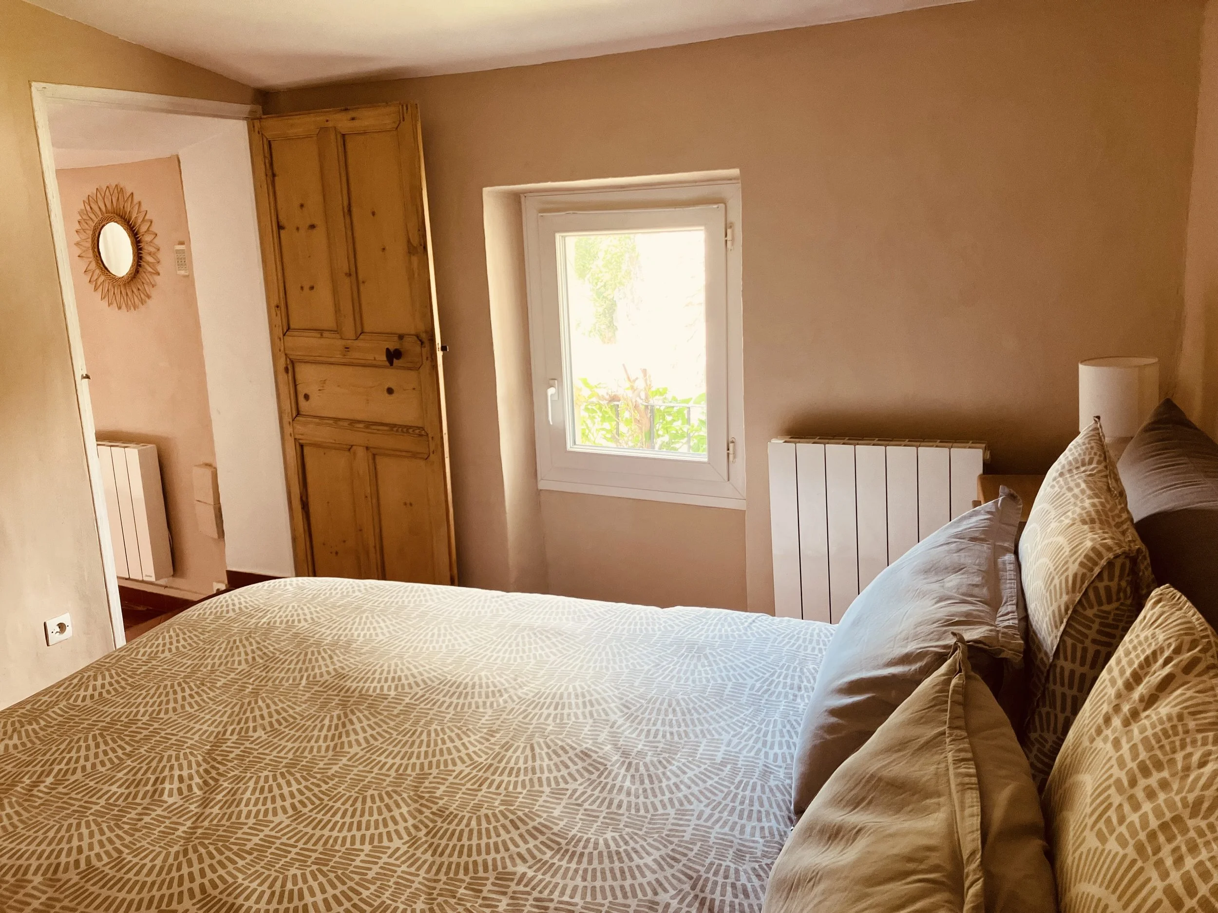 A bedroom with a bed that has a patterned beige and white bedspread and matching pillows. There is a wooden wardrobe against the wall, a window letting in natural light, a white radiator below the window, a white wall heater, and a round mirror with 