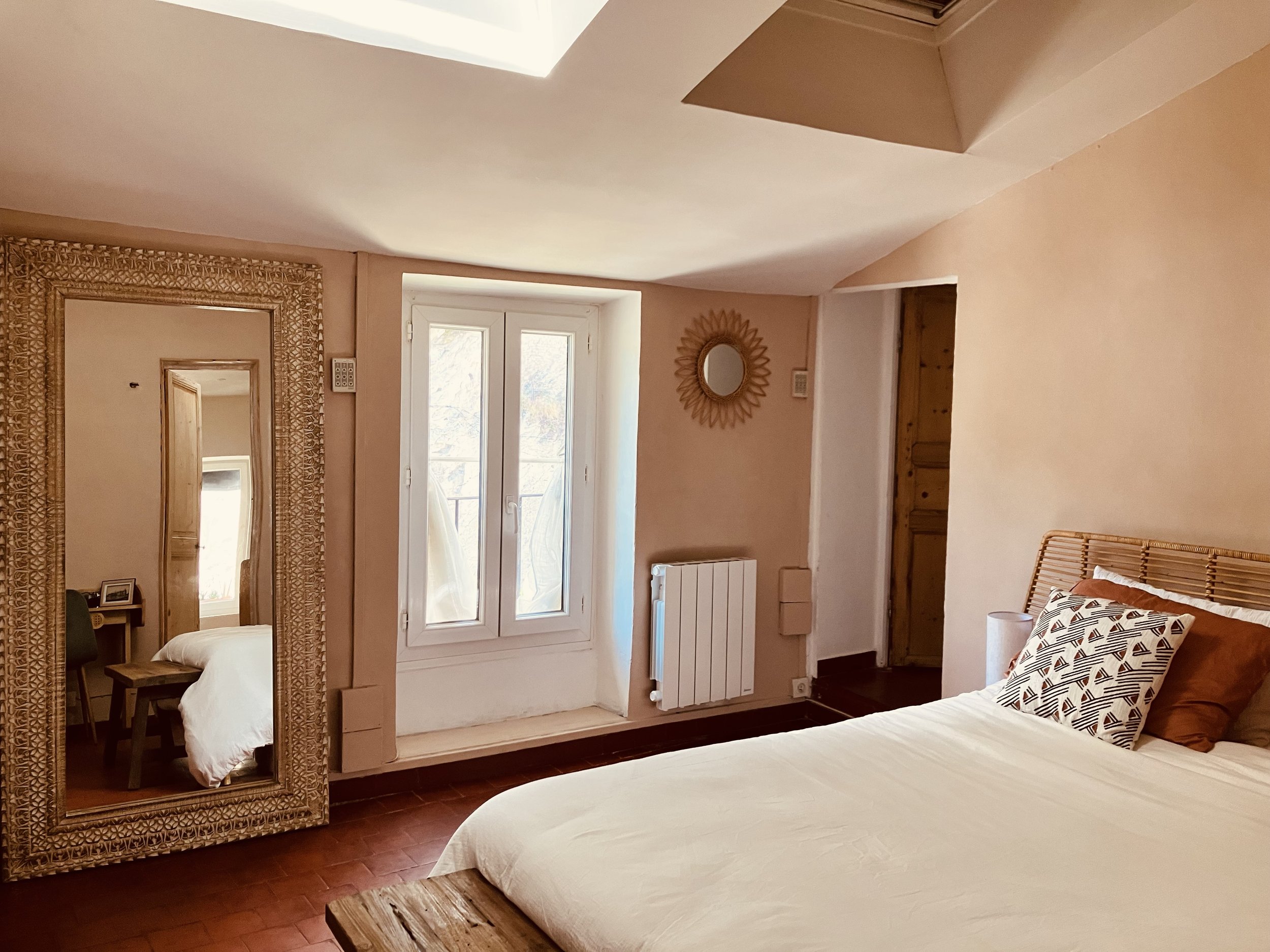 A cozy bedroom with a large window, a bed with white bedding, a wicker chair with patterned pillow, and decorative mirrors on the walls. The room has warm pink walls and wooden accents.