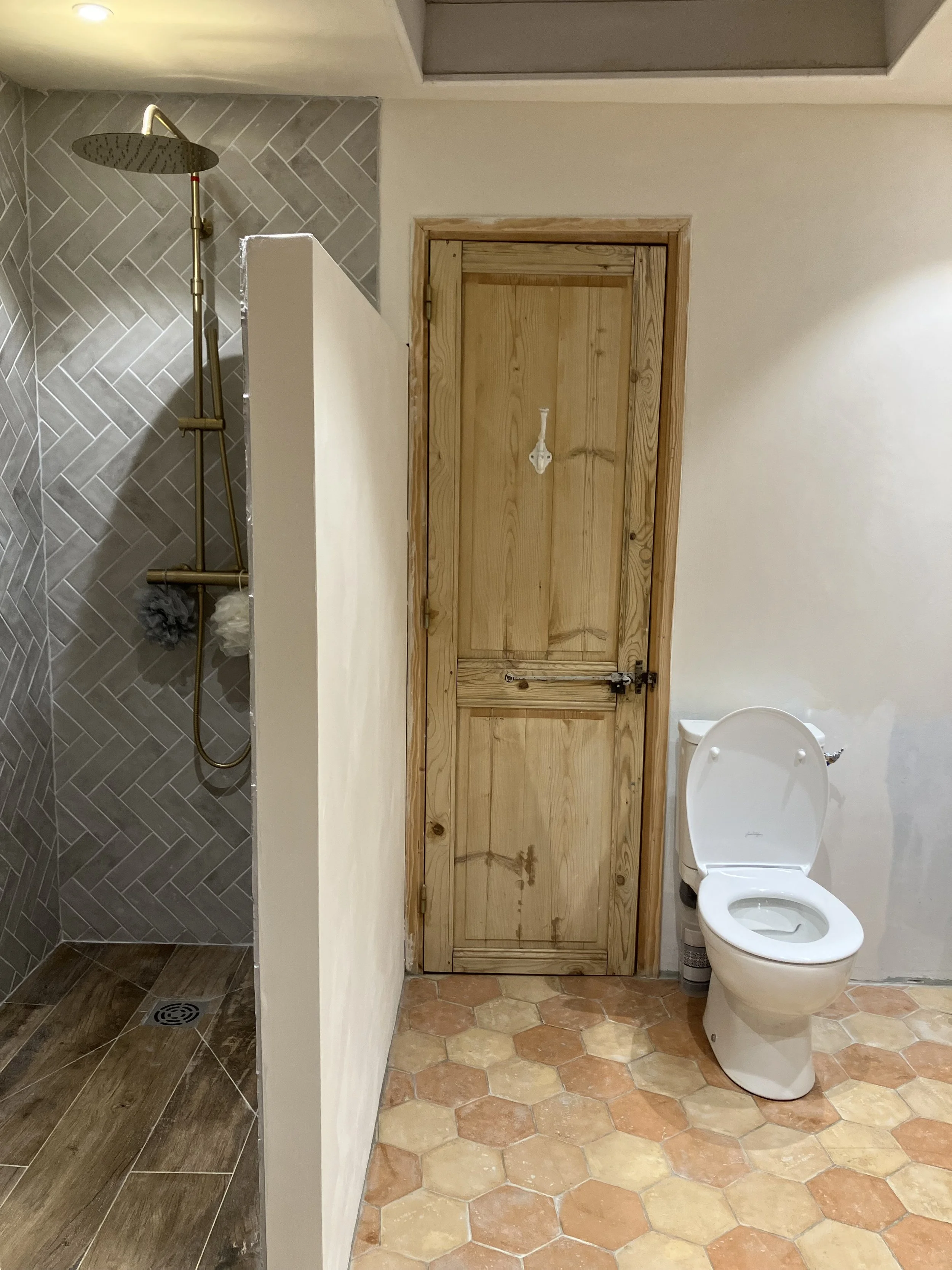 Bathroom with a shower area on the left with grey tile walls and brown wood-like floor tiles, separated by a partial wall from a toilet on the right, with white walls and a wooden door behind it.