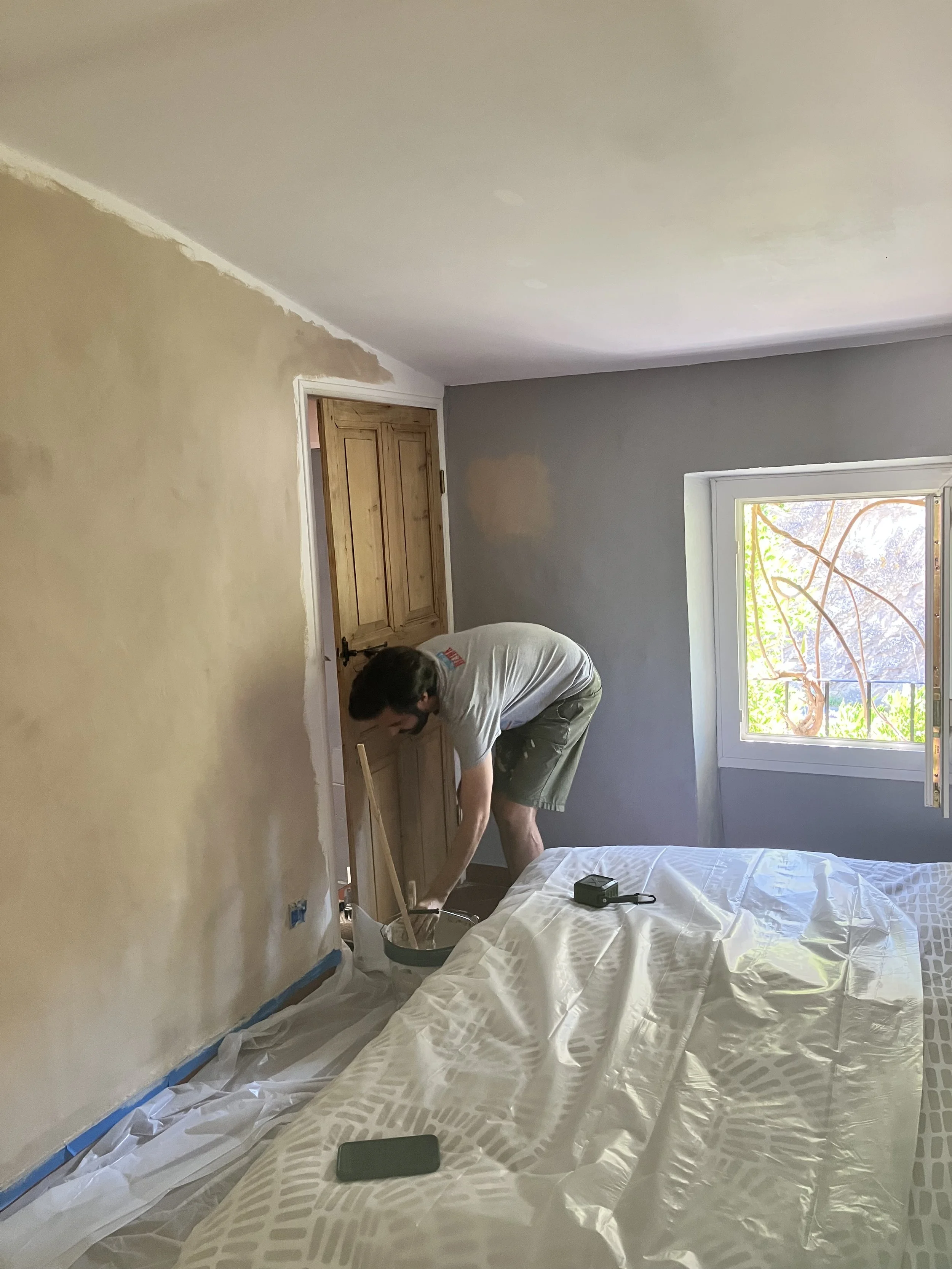 A man working on a home renovation project, painting or repairing a wooden door inside a room with a window.