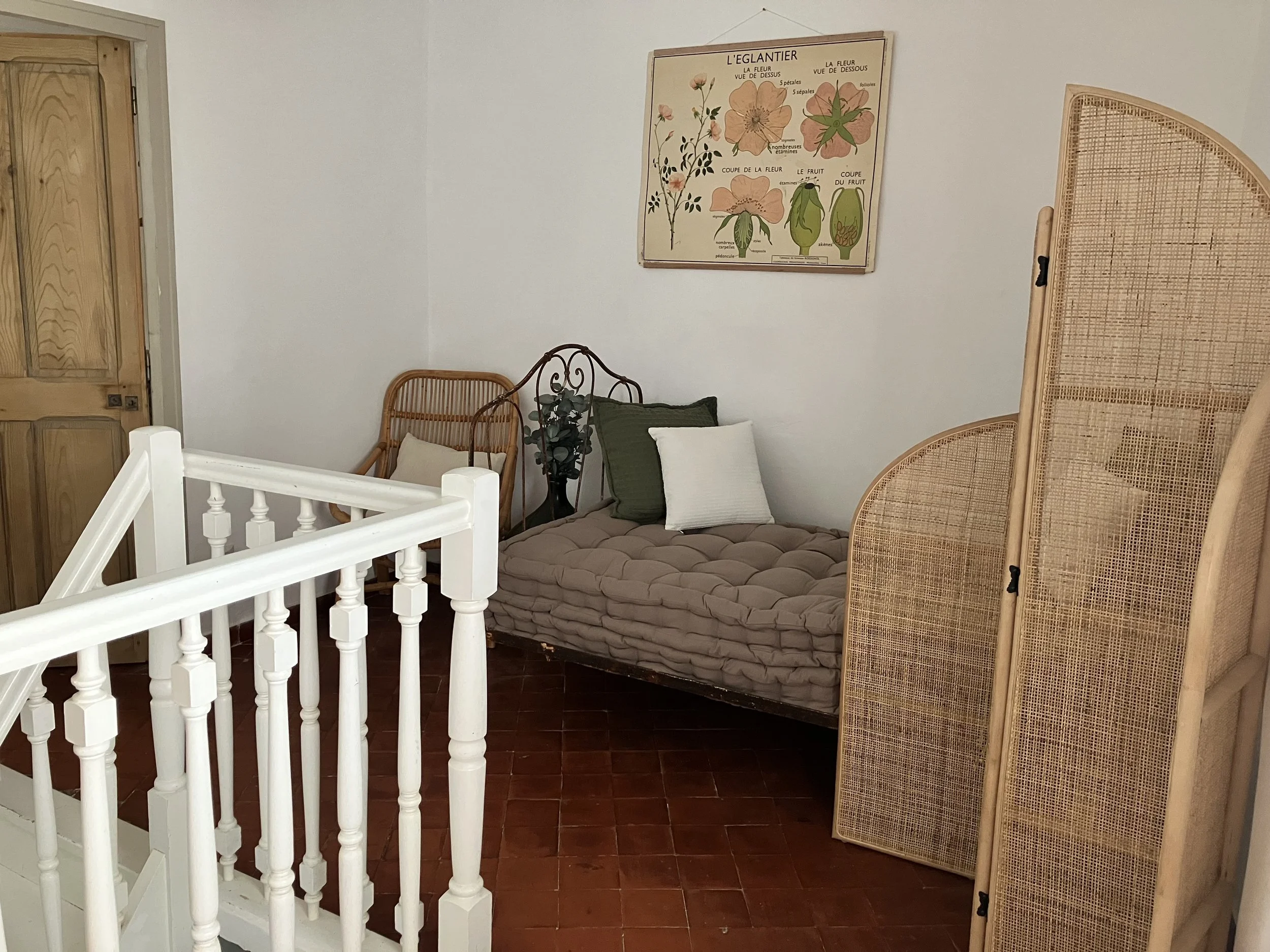 A cozy room with white walls and wooden flooring, featuring a bed with pillows, a wicker chair, a potted plant, and a decorative rattan folding screen. A framed botanical poster hangs on the wall.