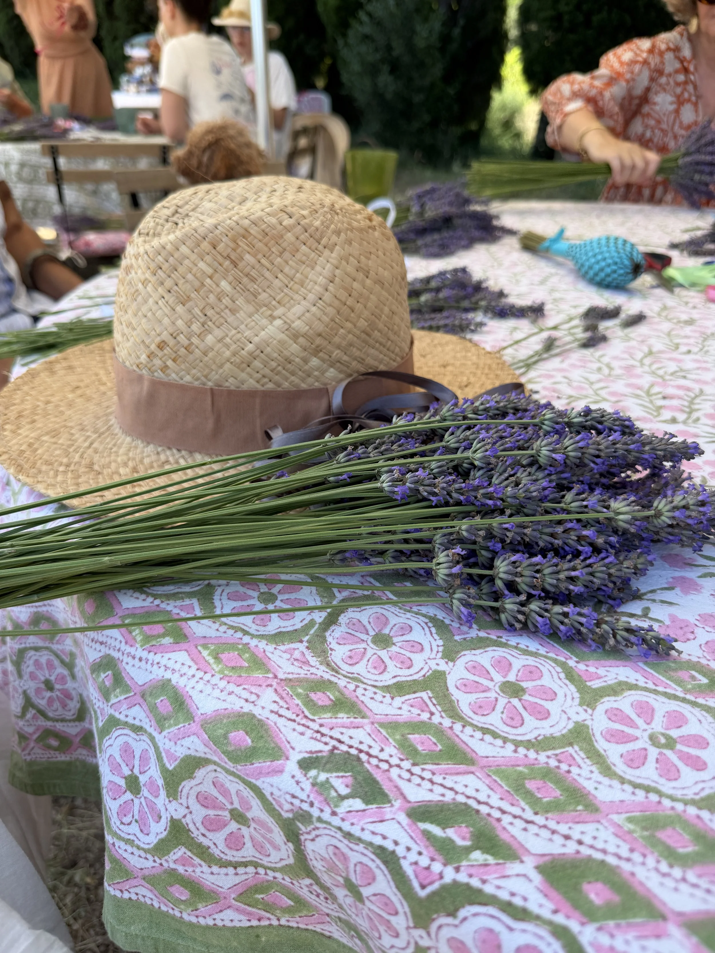 A table outdoors with a pink, green, and white patterned tablecloth, lavender flowers, a straw hat, and people in the background.