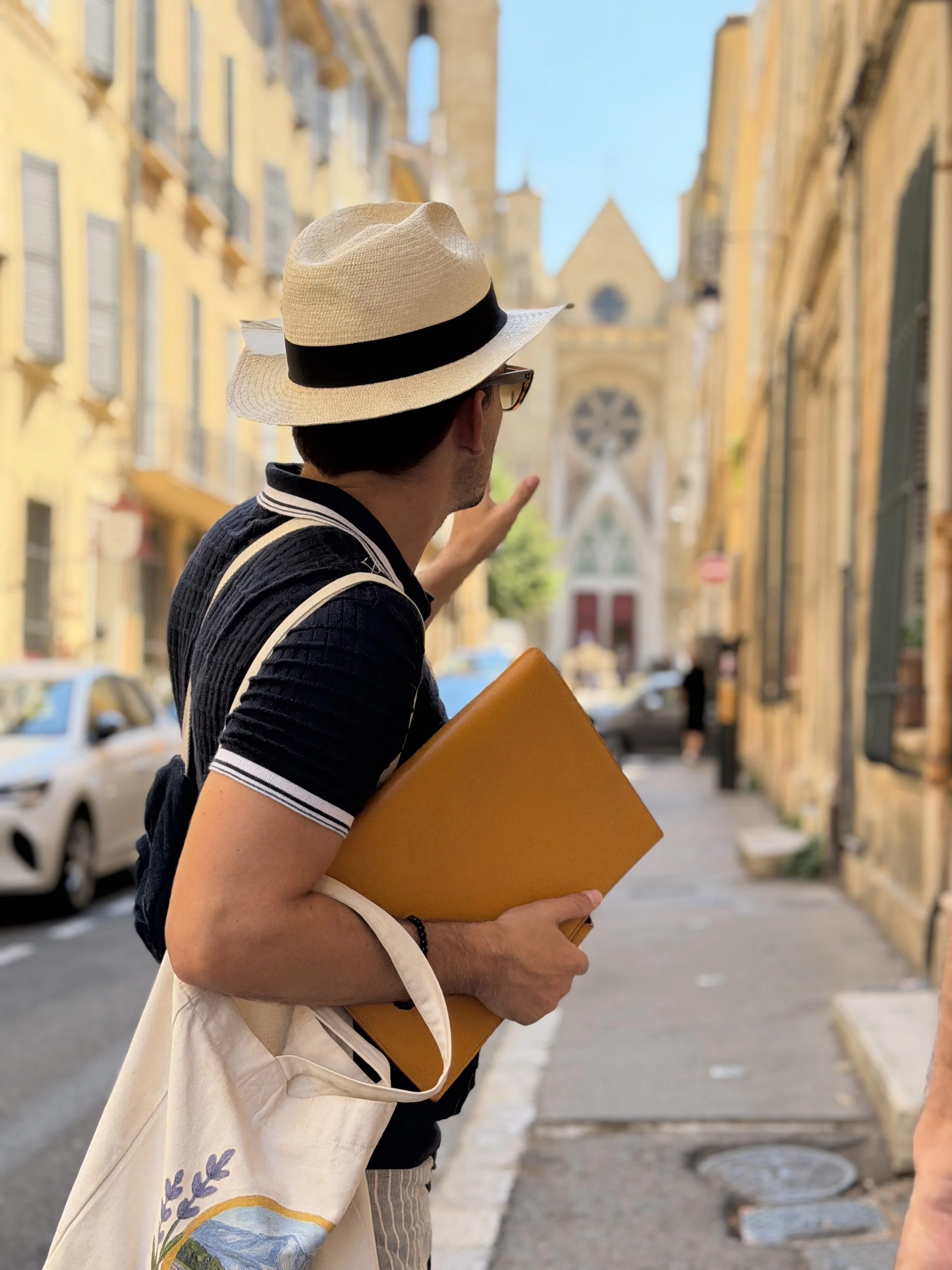 Man wearing a straw hat with a black band, sunglasses, a black shirt, and carrying a beige bag with a floral and landscape design, standing on a city street with yellow buildings and an church with a large circular window in the background.
