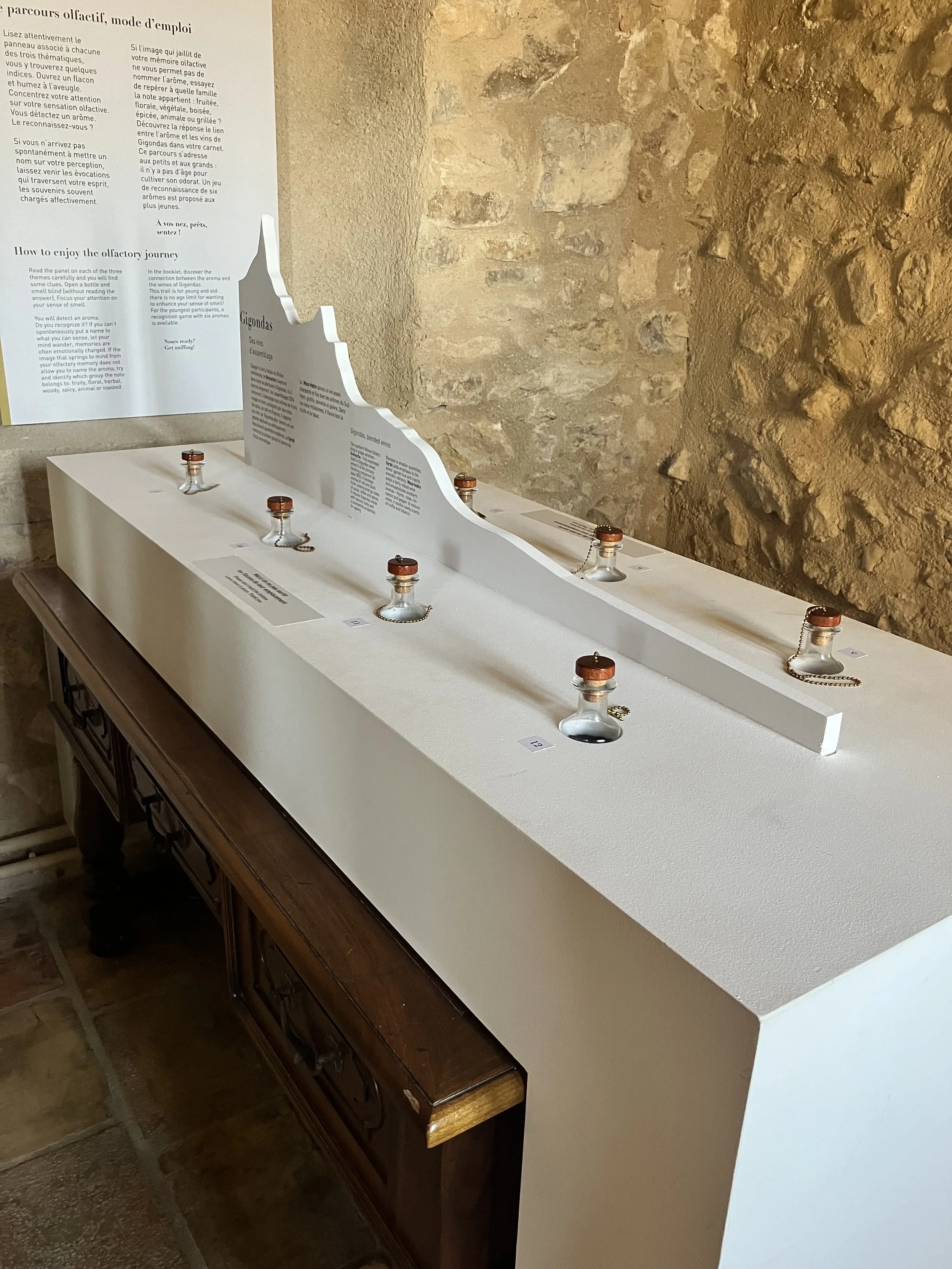 Museum display of small glass bottles with copper lids, chained to a white tabletop, with informational signs and a textured brick wall in the background.