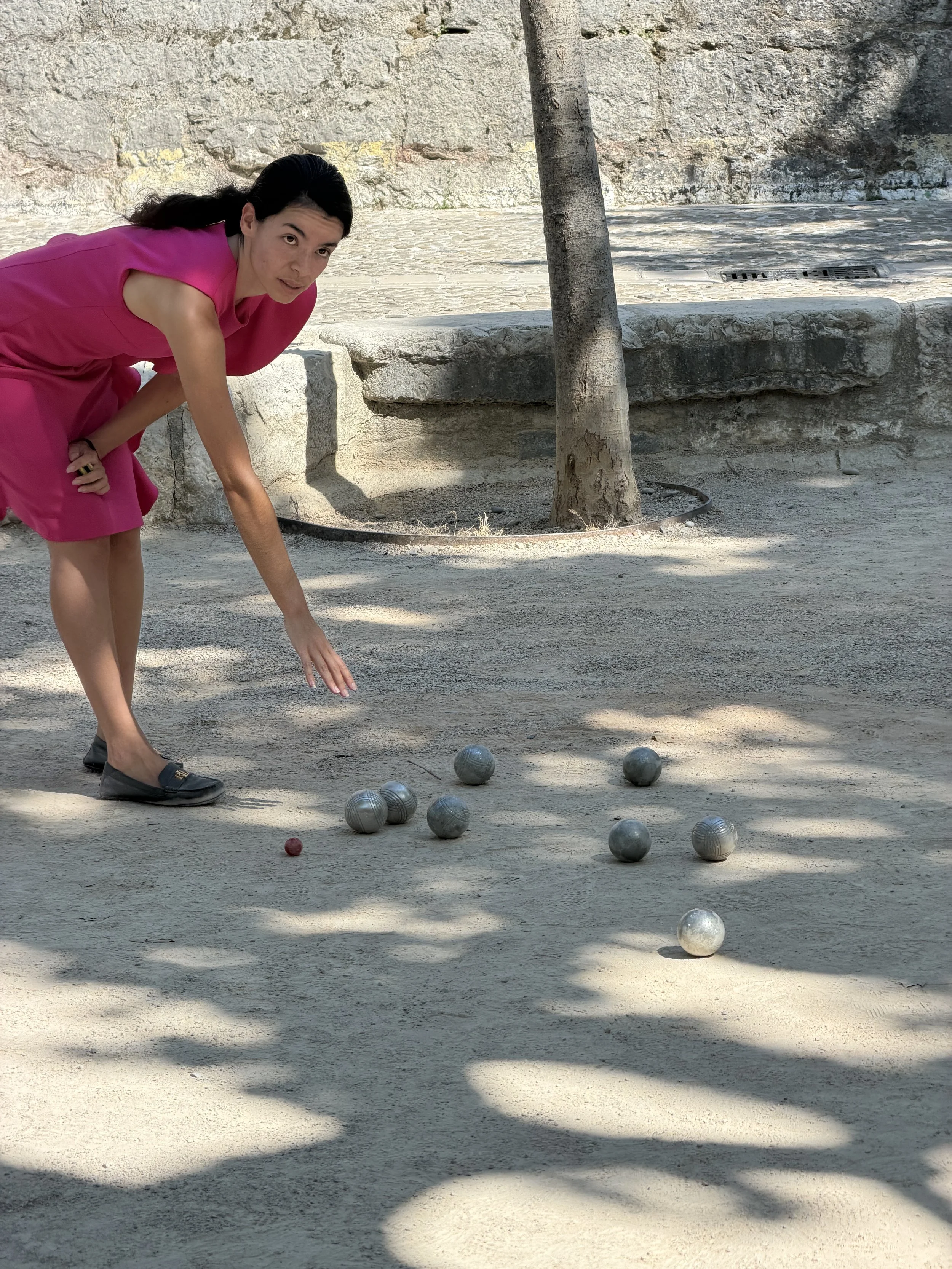 A woman in a pink dress playing pétanque outdoors, crouched down near silver metal balls and a red target ball, with a stone wall and tree in the background.