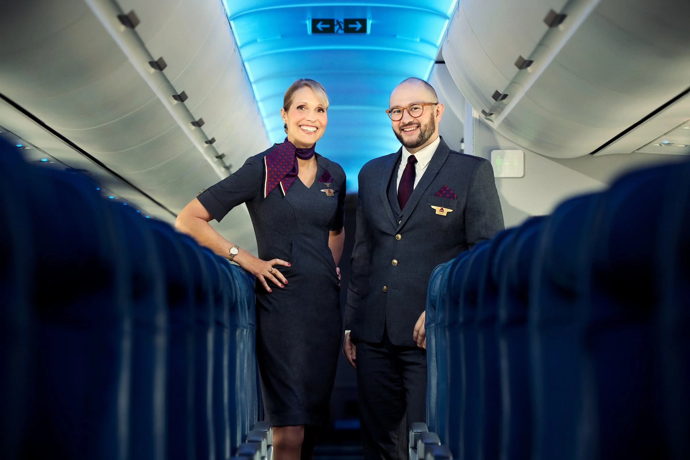 Two airline crew members, a woman and a man, standing in an airplane aisle smiling at the camera.