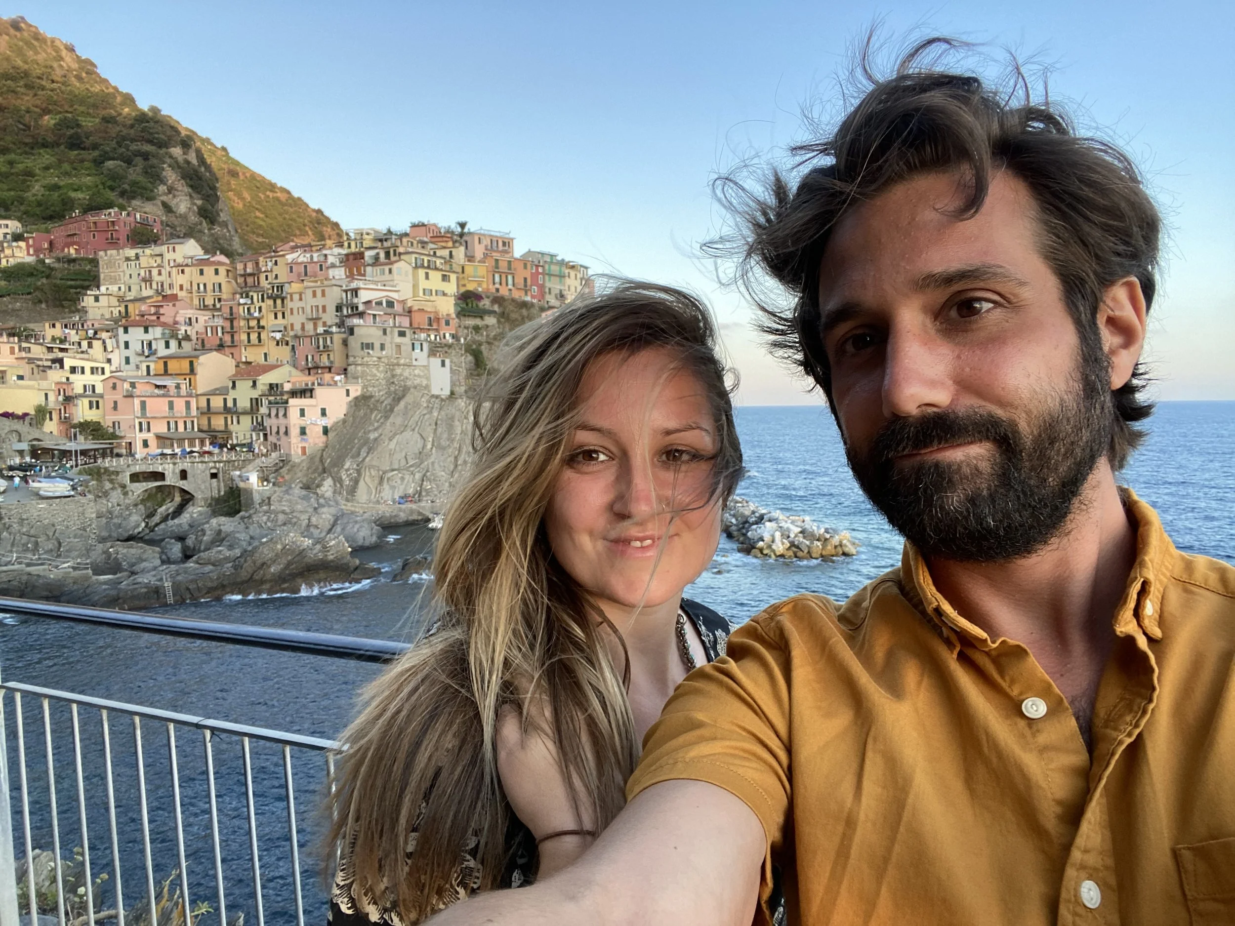 A man and woman taking a selfie on a bridge over the ocean with colorful buildings on a hillside in the background.