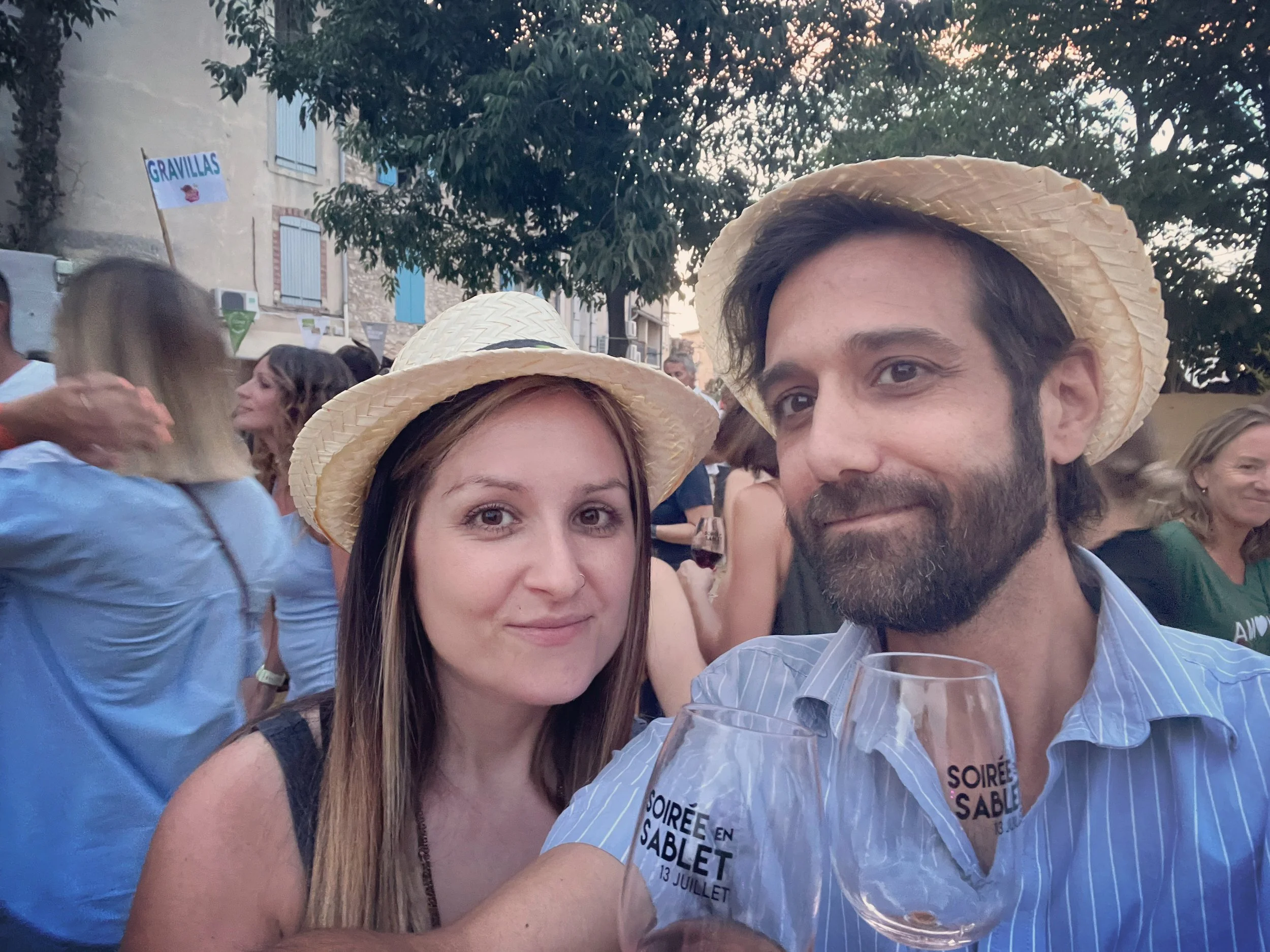 Close-up of a woman and a man wearing straw hats at a social gathering outdoors. The woman has long brown hair and the man has dark hair and a beard. They are surrounded by other people and trees, with a sign in the background reading 'GRAVILLAS'.