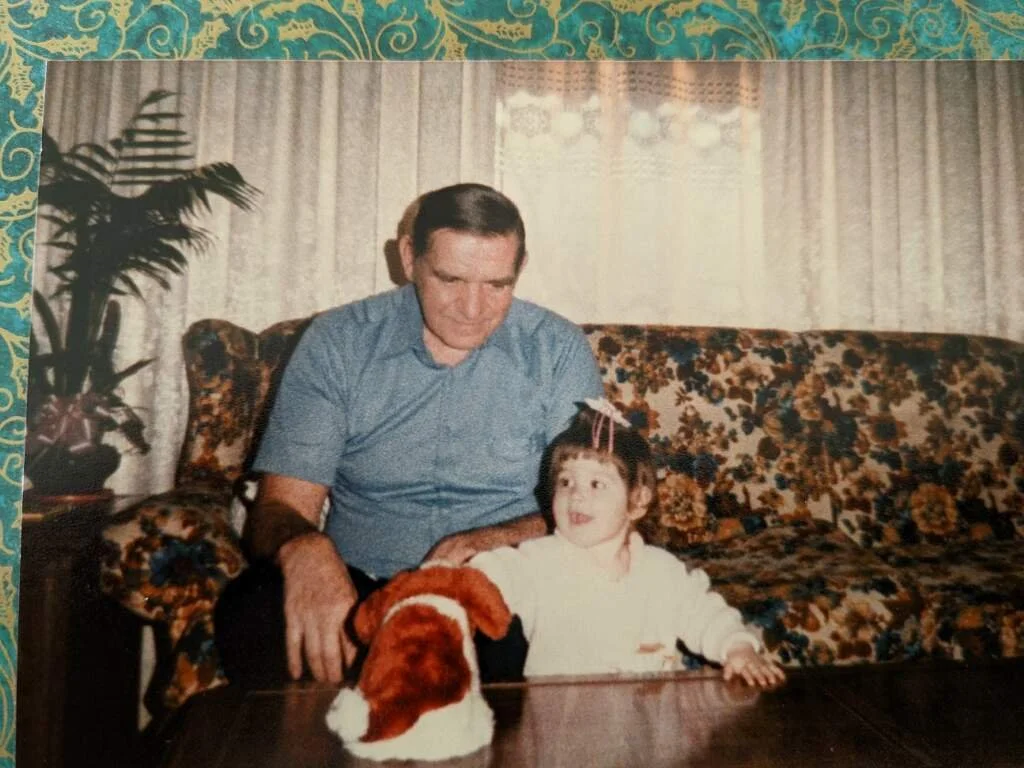 A young girl sitting on a floral patterned sofa with an older man beside her, a stuffed dog toy on the table, and a potted plant in the background in a living room.