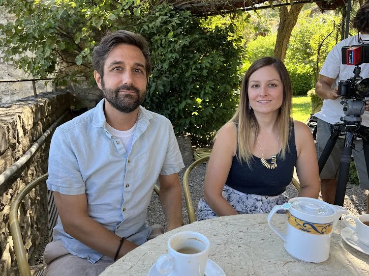 A man with a beard and a woman with long blonde hair sitting at an outdoor table. There is a camera operator in the background.