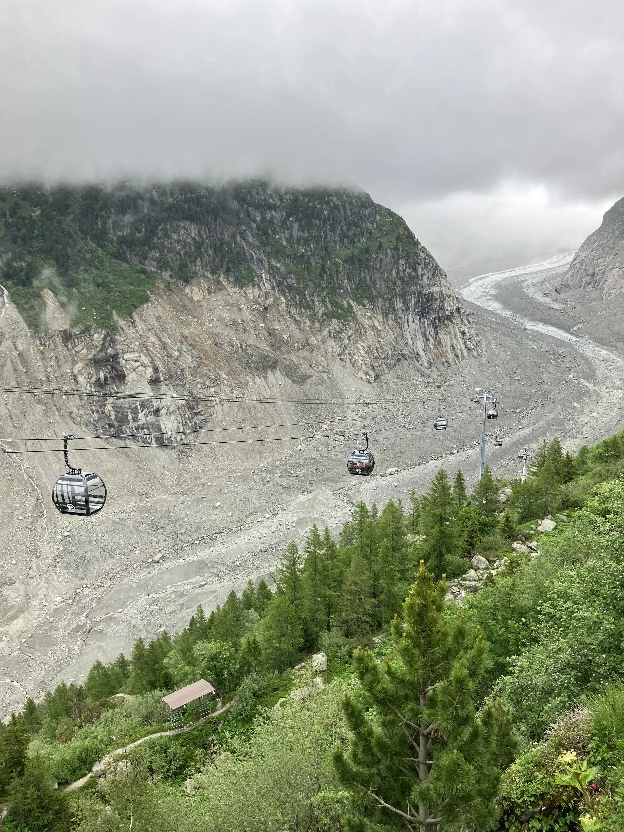 A mountain landscape with green forests, rocky slopes, and a glacier in the background. Cable cars are visible in the foreground, traveling over rocky terrain with a small shelter at the bottom.