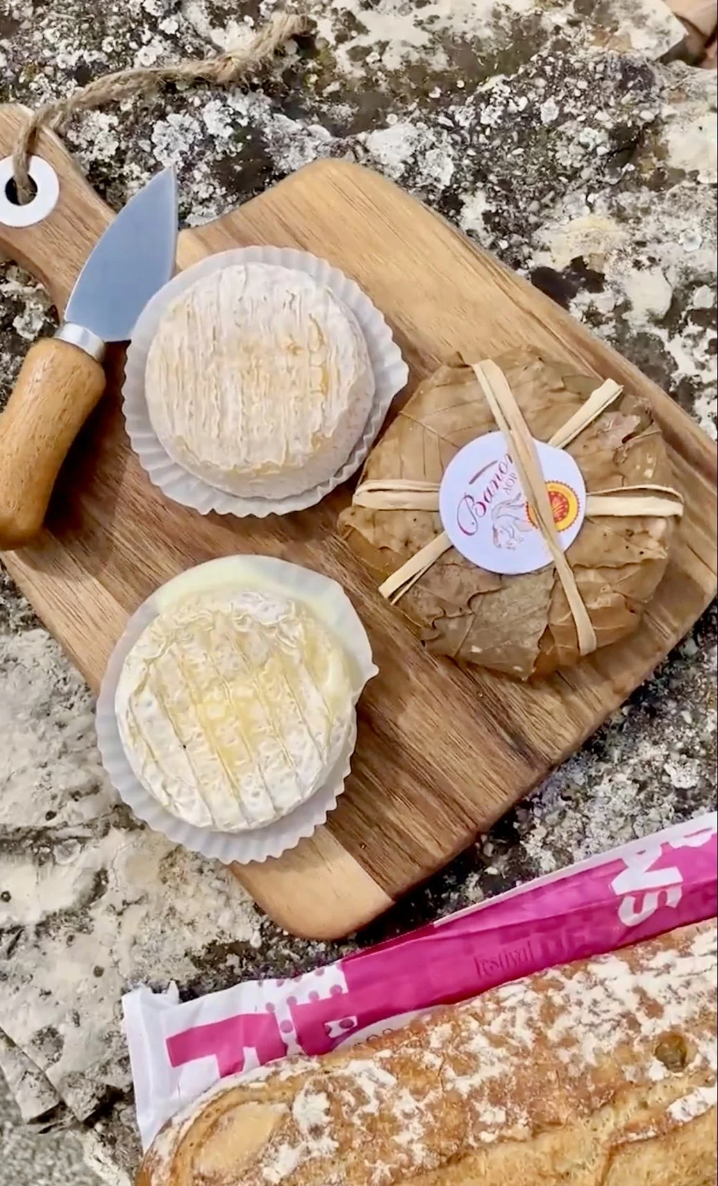 Two wheels of soft cheese on parchment paper, a wrapped and labeled cheese, and a bread loaf on a wooden cutting board outdoors.