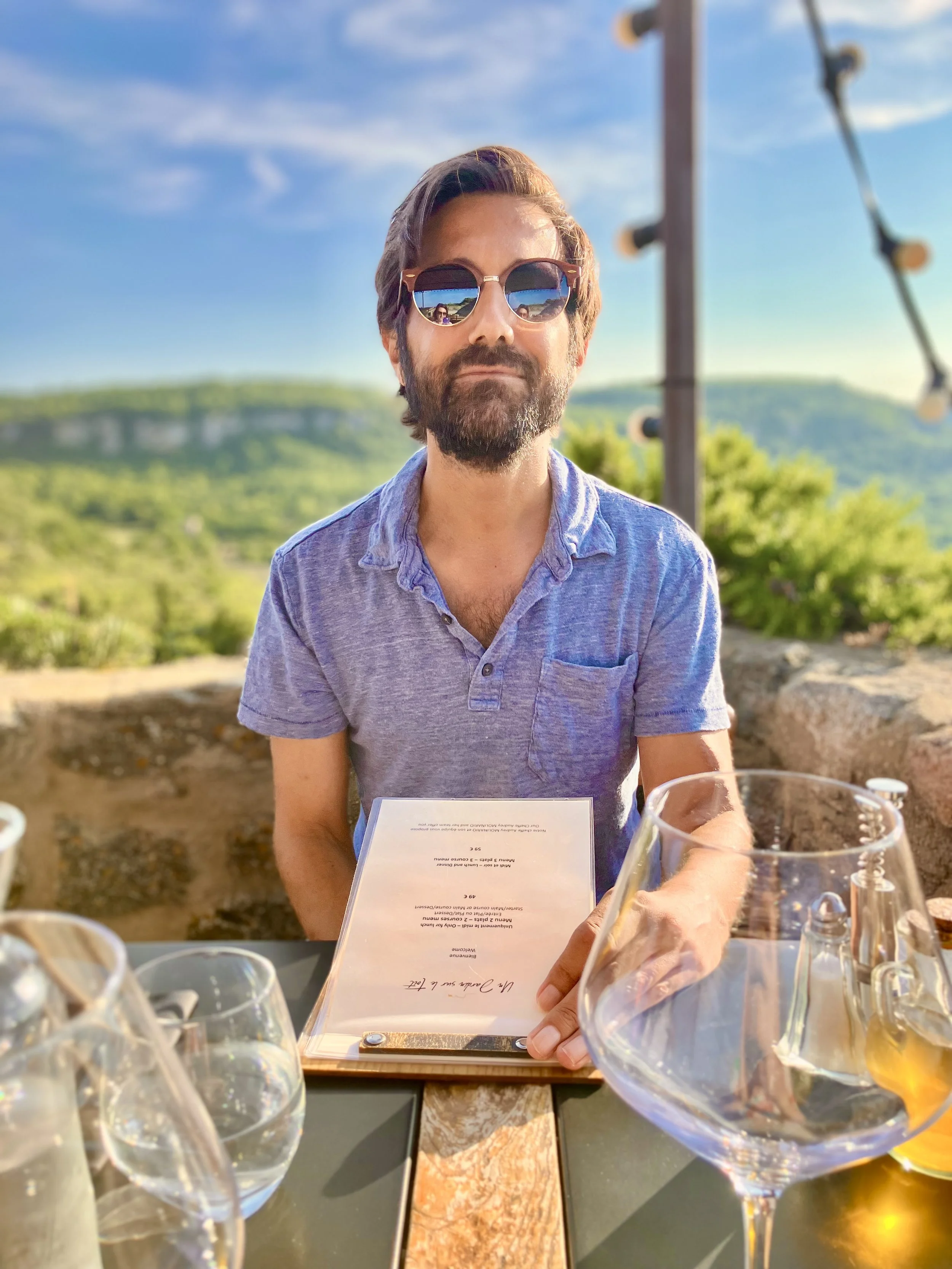 A man with glasses and a beard sitting at an outdoor restaurant table with a menu in front of him, few glasses of wine and water on the table, a person reaching with a hand on the right side, and a scenic view of green hills and blue sky in the background.