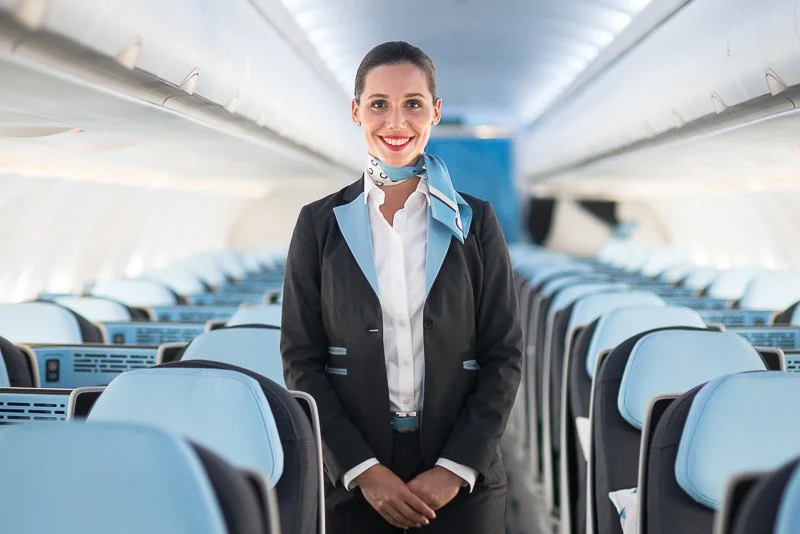 A smiling female flight attendant standing inside an airplane cabin.