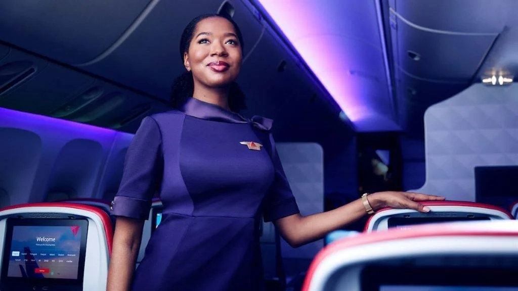 A flight attendant standing inside an airplane cabin, smiling, with her hand resting on a seat. The cabin is illuminated with blue and purple lighting.