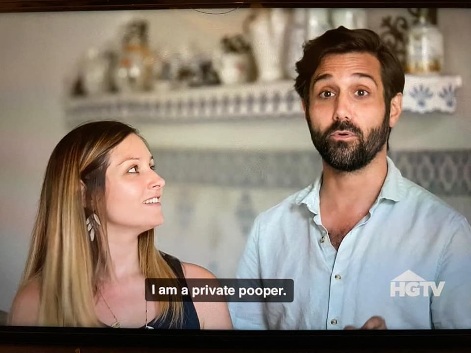 A man and woman having a conversation indoors. The man has dark hair and a beard, and is wearing a light blue shirt. The woman has long, light brown hair and is looking at the man. The screen displays the text, "I am a private pooper." The HGTV logo is in the bottom right corner.