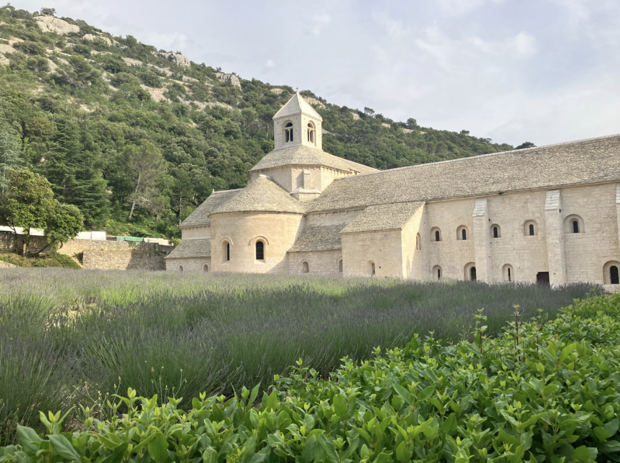 A historic stone church with a bell tower, surrounded by lush green mountains and a garden of lavender and greenery in the foreground.
