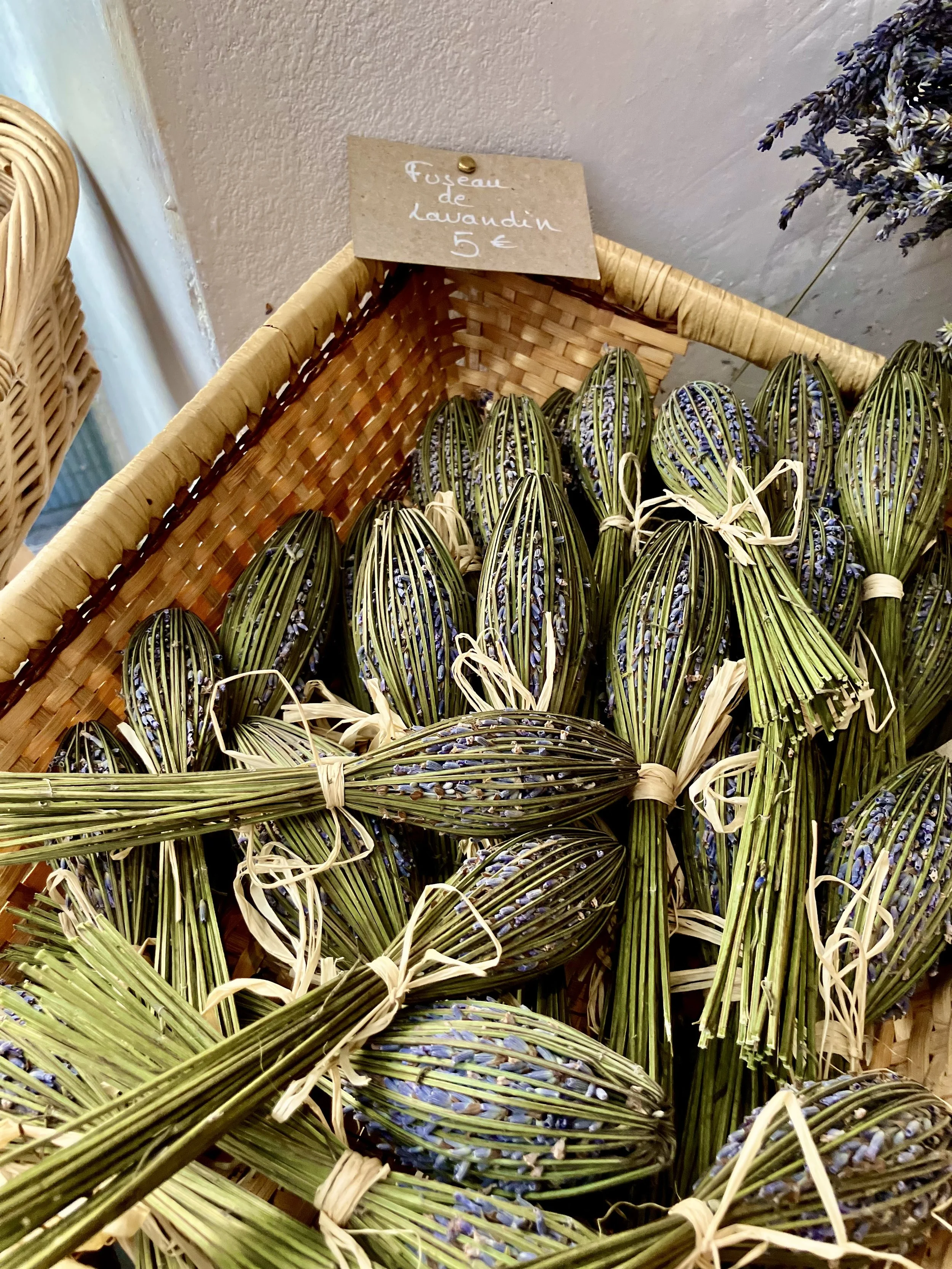 Bundles of lavender flowers tied together and displayed in a basket with a sign that reads 'Fuseau de lavandin 5 €'.