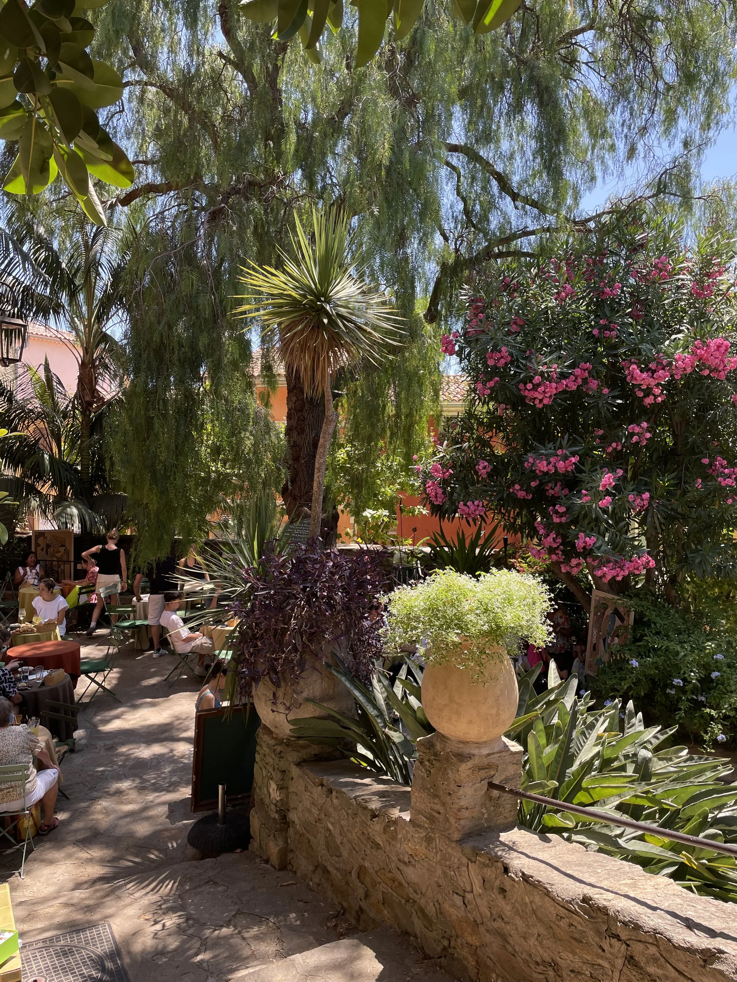 A lively outdoor patio surrounded by lush green trees and flowering bushes, with several people dining at tables under the shade on a sunny day.