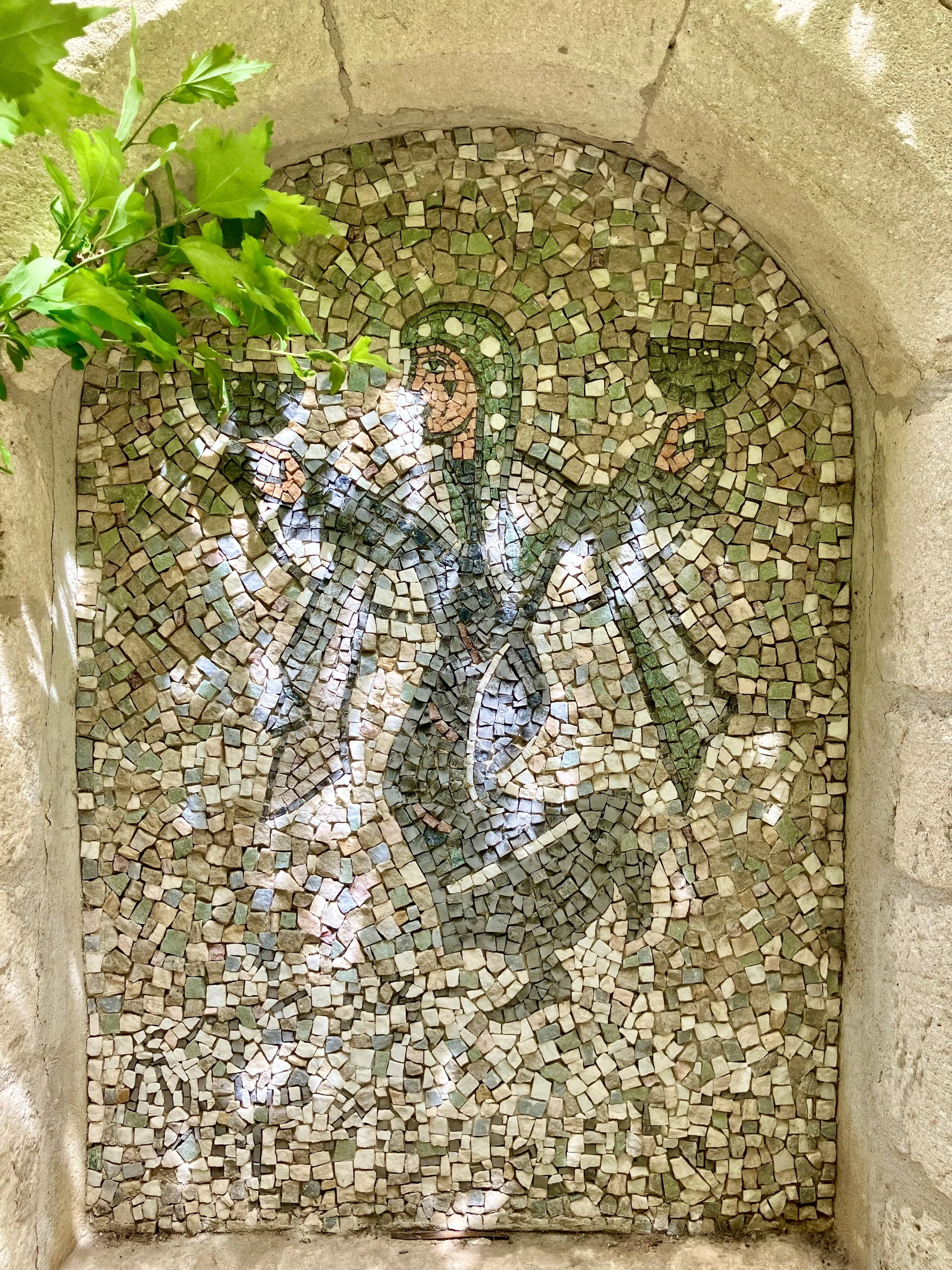 A mosaic art piece with a floral and bird design, surrounded by a stone wall and some green leaves in the upper left corner.