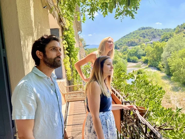Three people standing on a balcony overlooking a lush, green river valley on a sunny day.