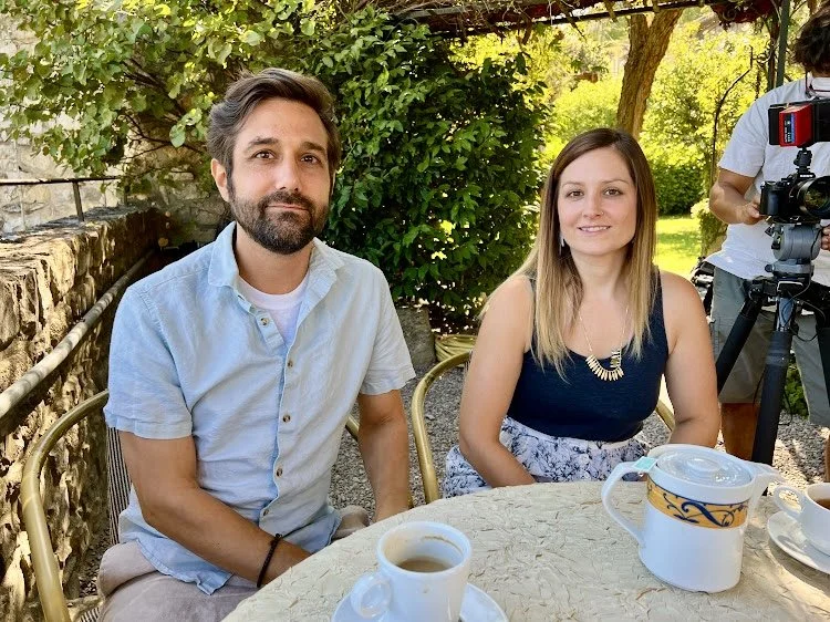 A man with a beard and a woman sitting at an outdoor table with coffee cups, with a person filming in the background and greenery around.