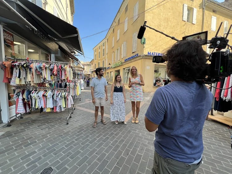 A camera operator filming three people on a street with yellow buildings, clothing stalls, and clear blue sky.