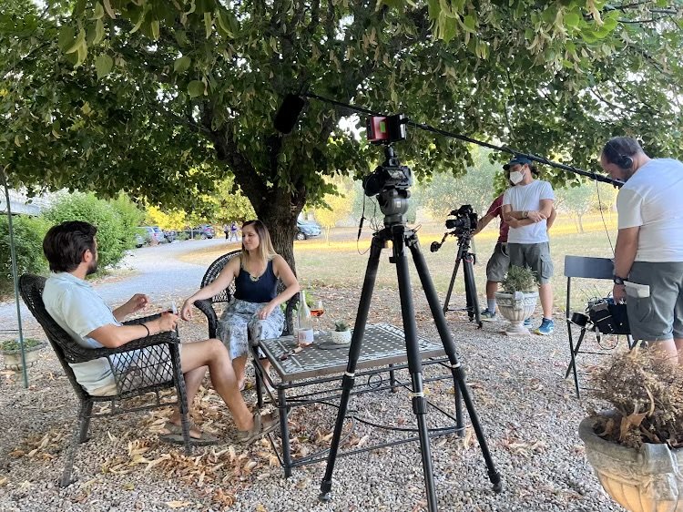 Filming crew conducting an outdoor interview under a large tree, with two people sitting and talking at a small table, surrounded by filming equipment.