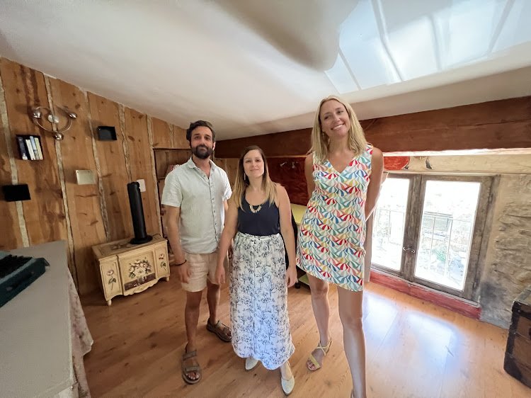 Three people standing inside a room with wooden walls and a large window. Two women and one man, all smiling at the camera. The woman on the right is wearing a colorful dress, and the woman in the middle is wearing a navy top with a white skirt. The man on the left is wearing a white shirt and beige shorts.