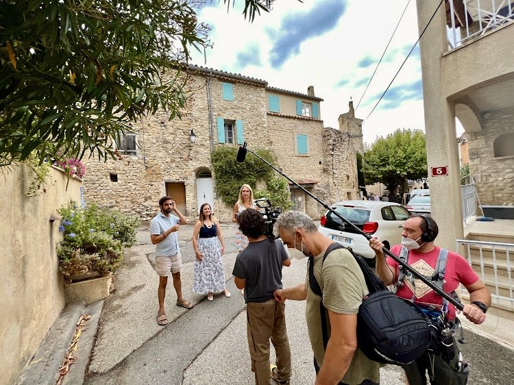 A film crew filming an interview or scene with three women standing in a street in front of historic buildings. The crew includes a camera operator, a sound technician, and a director. The weather is partly cloudy.