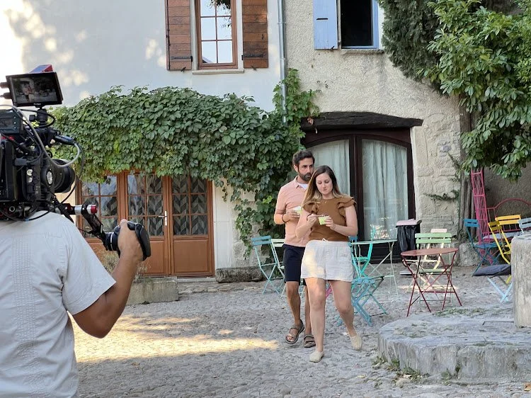 A man and woman walking on cobblestone street in front of a house with shutters and a large ivy plant, as a cameraman films them.