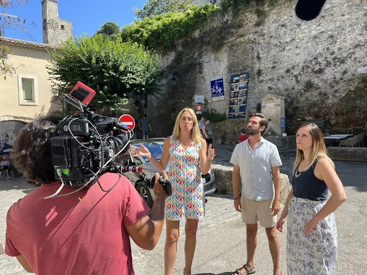 A woman in a colorful dress is speaking to a camera crew with a camcorder, while two people stand nearby listening, outdoors in a sunny location near a stone wall and trees.