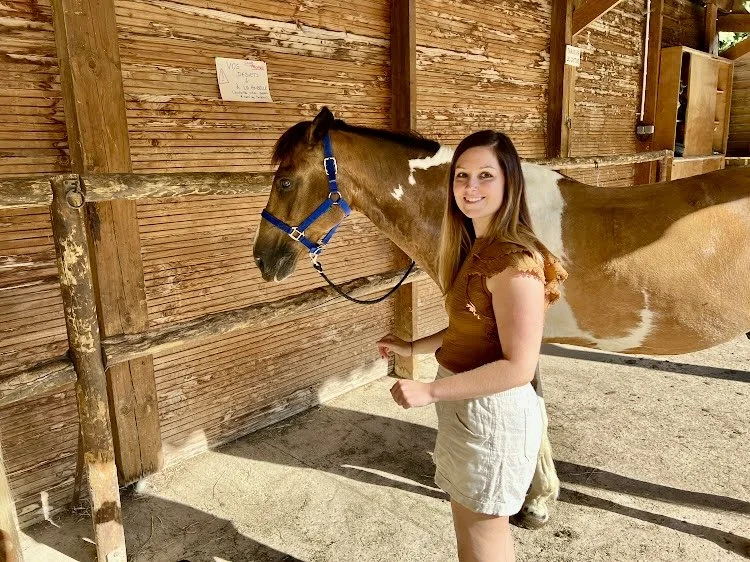 A woman standing next to a brown and white horse inside a wooden stable, smiling at the camera.