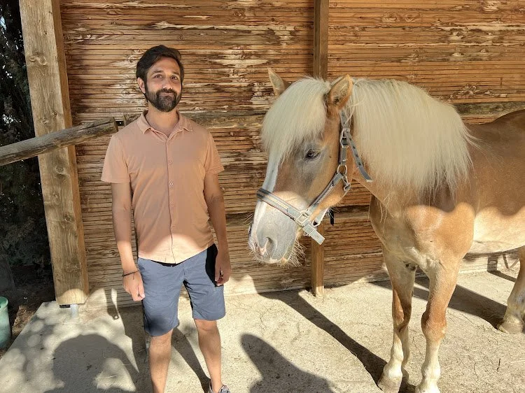 A man with a beard and mustache, wearing a peach-colored shirt and blue shorts, standing next to a tan horse with a light blonde mane inside a wooden stable.