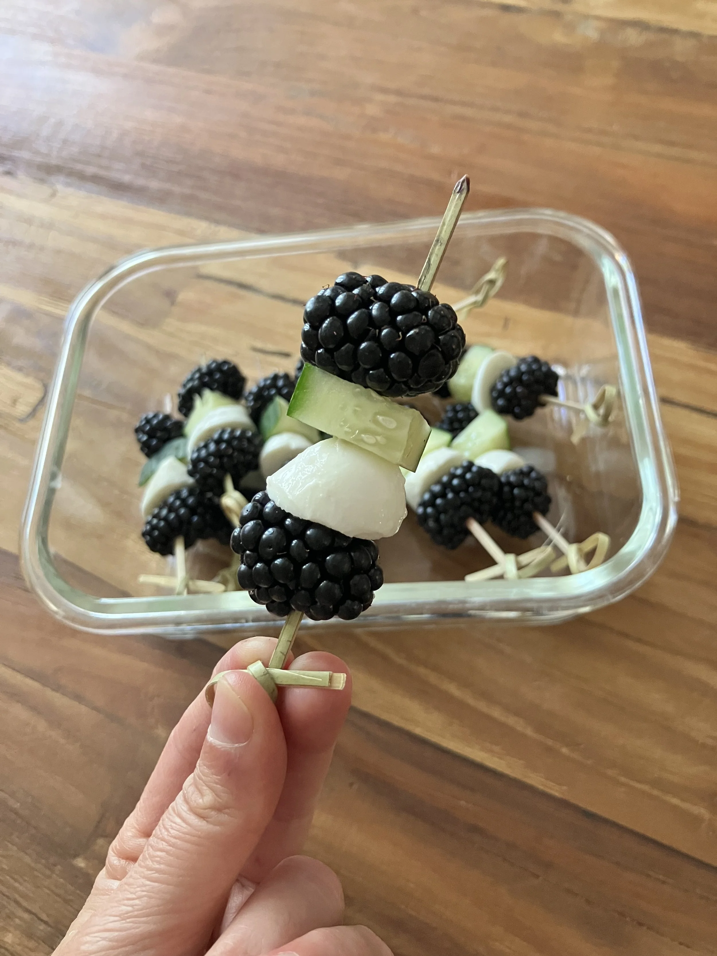 Close-up of a hand holding a skewer with blackberries, cucumber slices, and cheese. The background shows a wooden surface and a glass container filled with similar skewers.