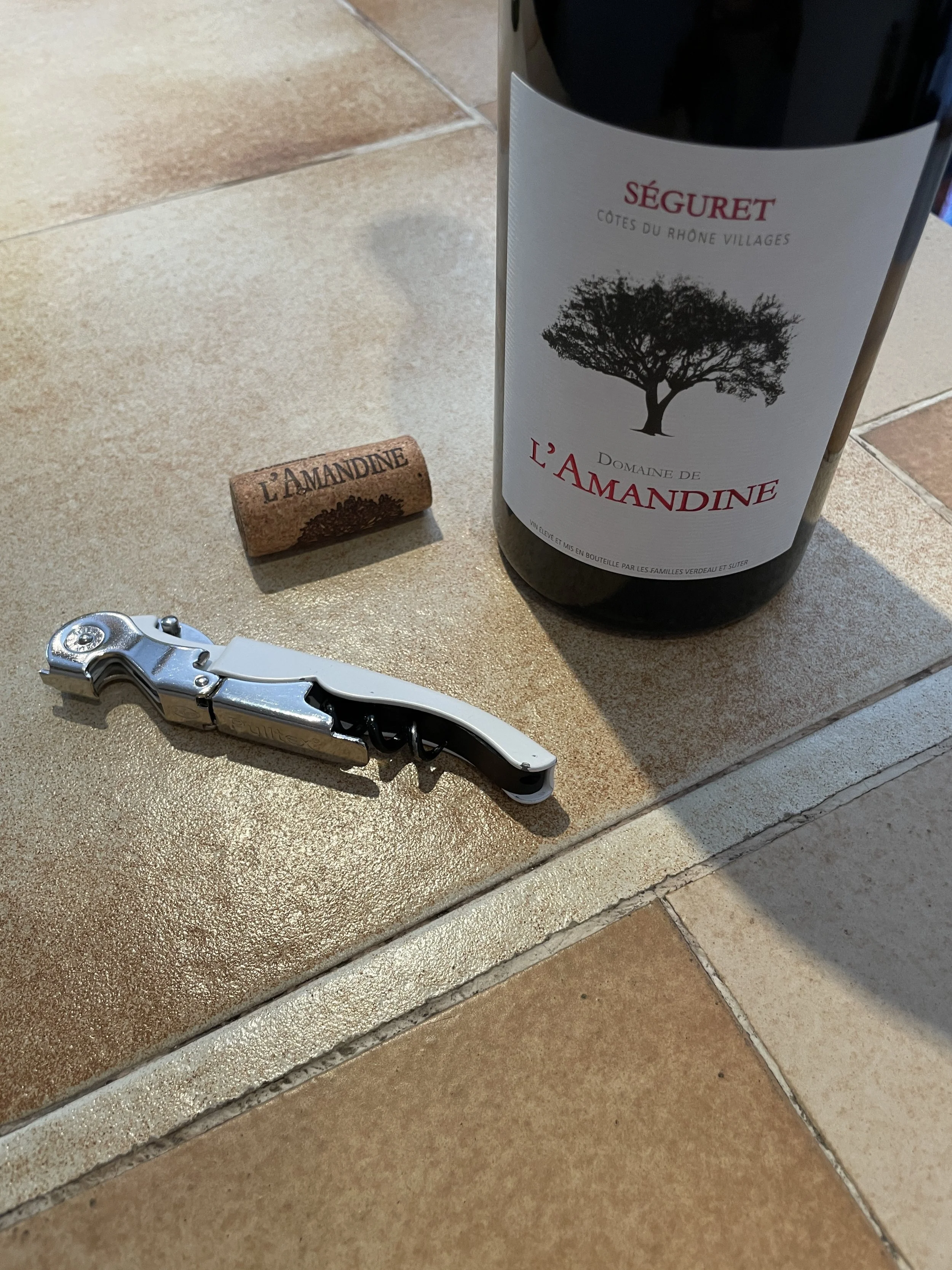 A bottle of red wine labeled 'Seguret L'Amadine', a wine cork with the same name, and a metal corkscrew on a tiled countertop.