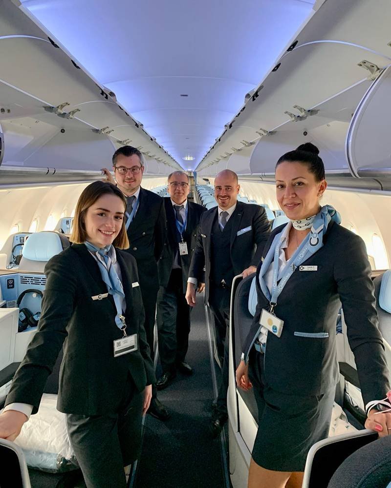 Five airline crew members standing inside an airplane cabin, smiling at the camera.