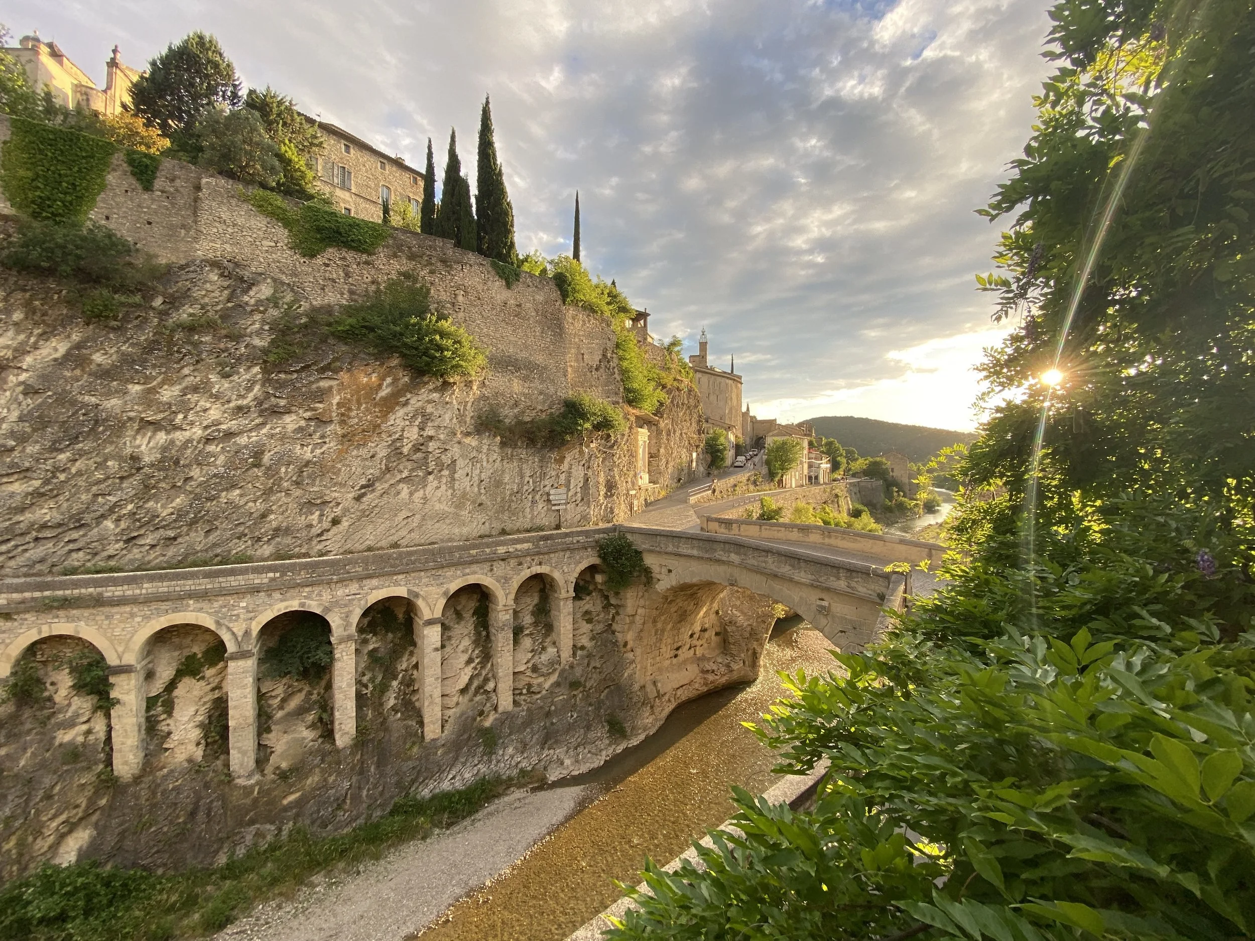 A historic stone building with tall cypress trees on a hill, a stone bridge over a river, and a winding road in a scenic town at sunset