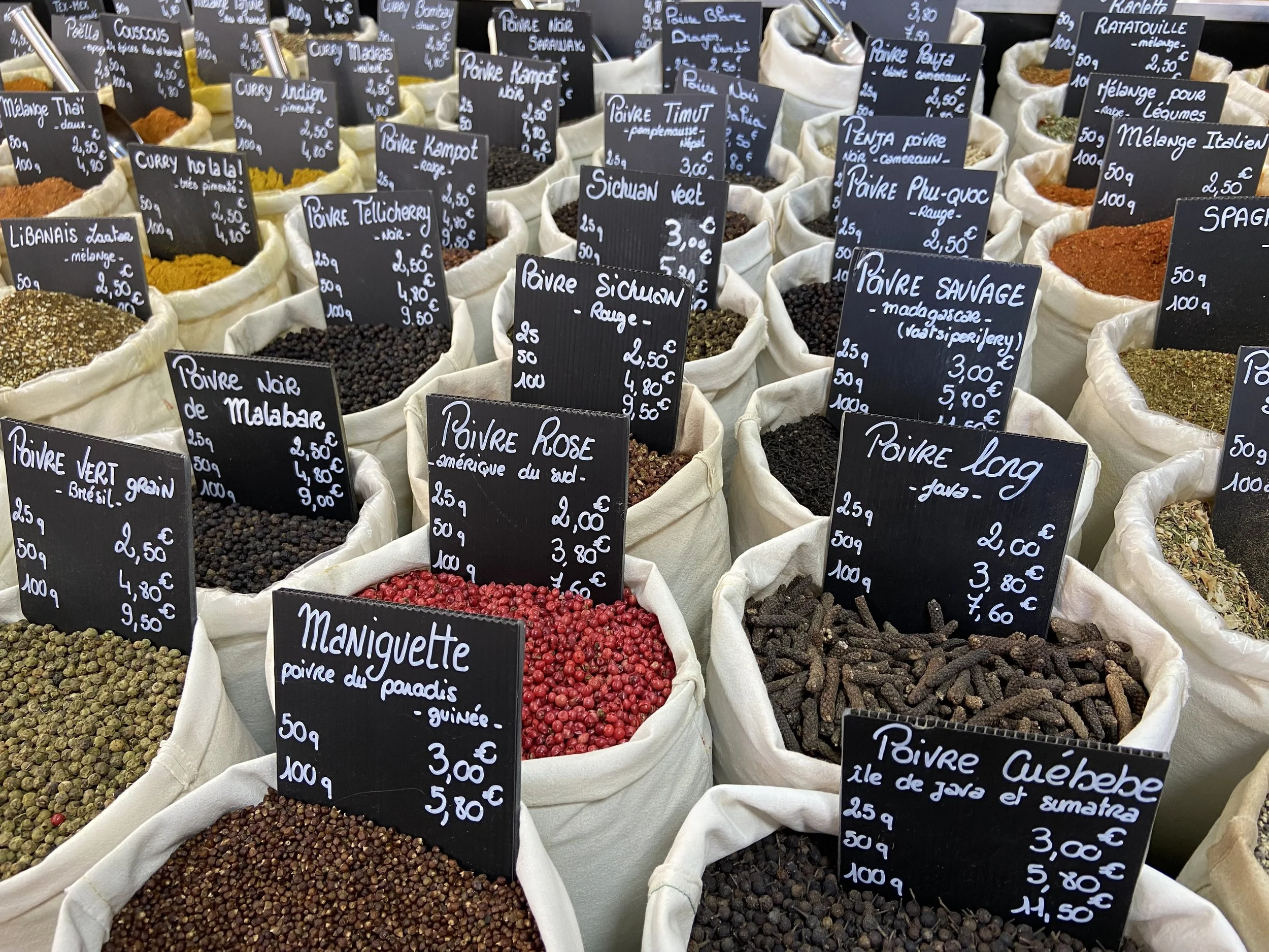 Various spices and herbs in cloth bags at a market, with small black signs displaying their names and prices in French.