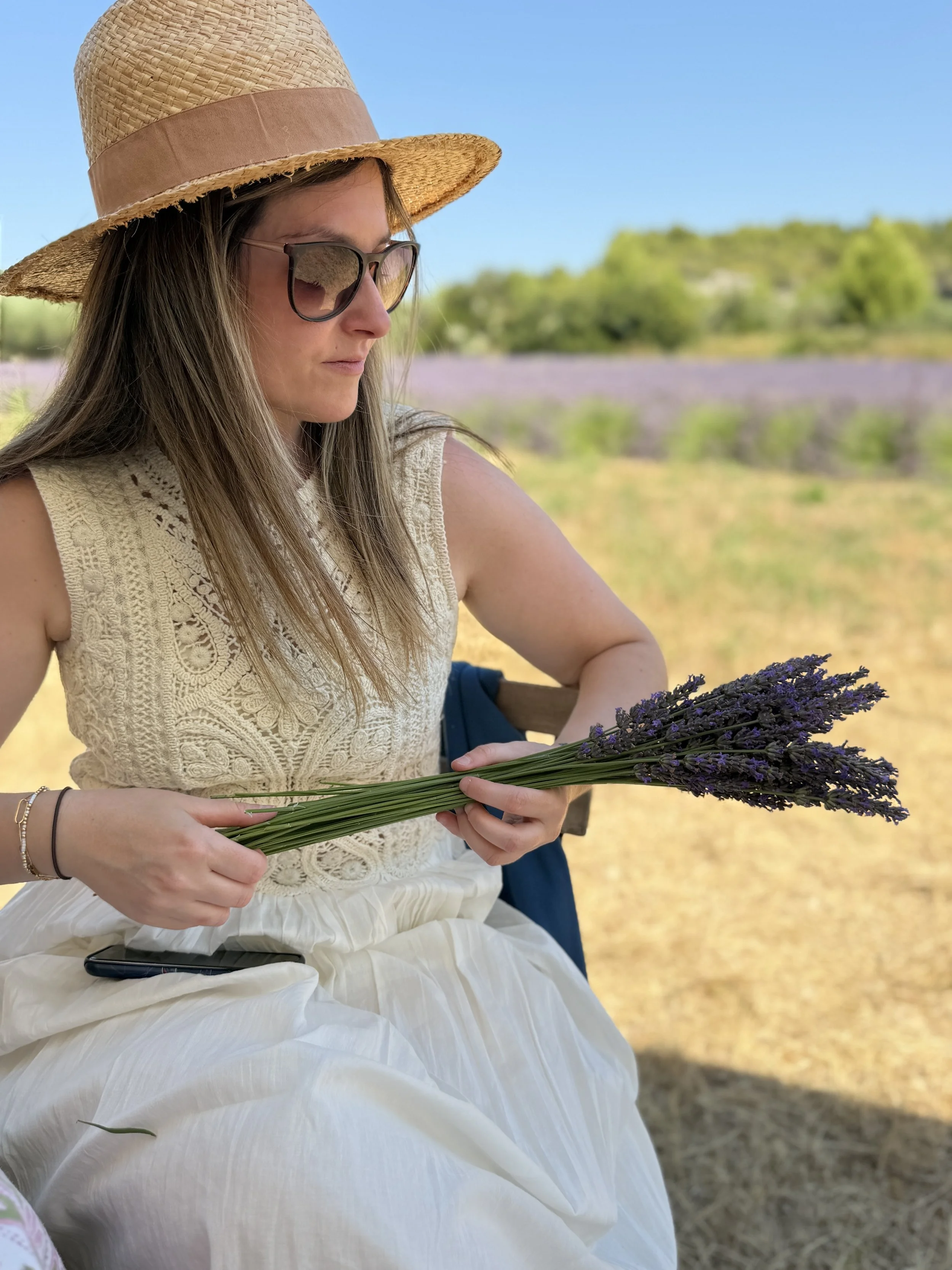 A woman wearing a straw sun hat, sunglasses, a cream-colored crochet top, and a white skirt, sitting outdoors on a sunny day, holding a bunch of purple lavender flowers.