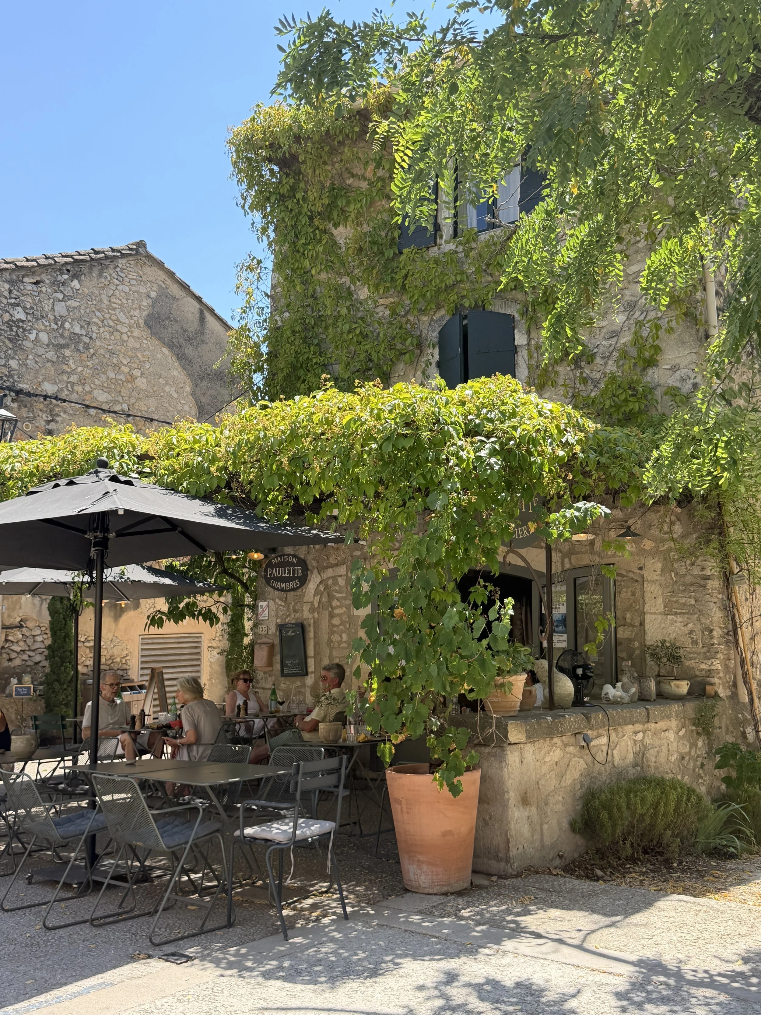 Outdoor cafe scene with several people sitting at tables under umbrellas, surrounded by greenery and stone buildings, with bright sunlight and clear blue sky.