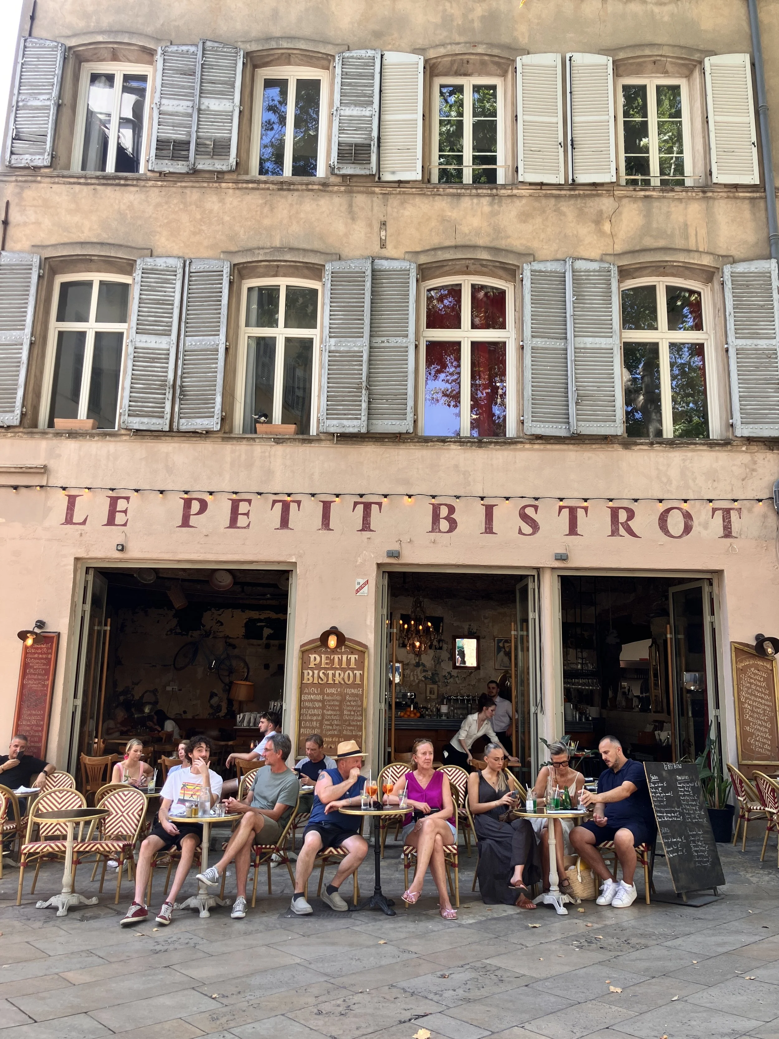Outdoor seating area of a restaurant called Le Petit Bistrot with people dining and chatting, while waitstaff serve food in front of a building with multiple windows and open shutters.