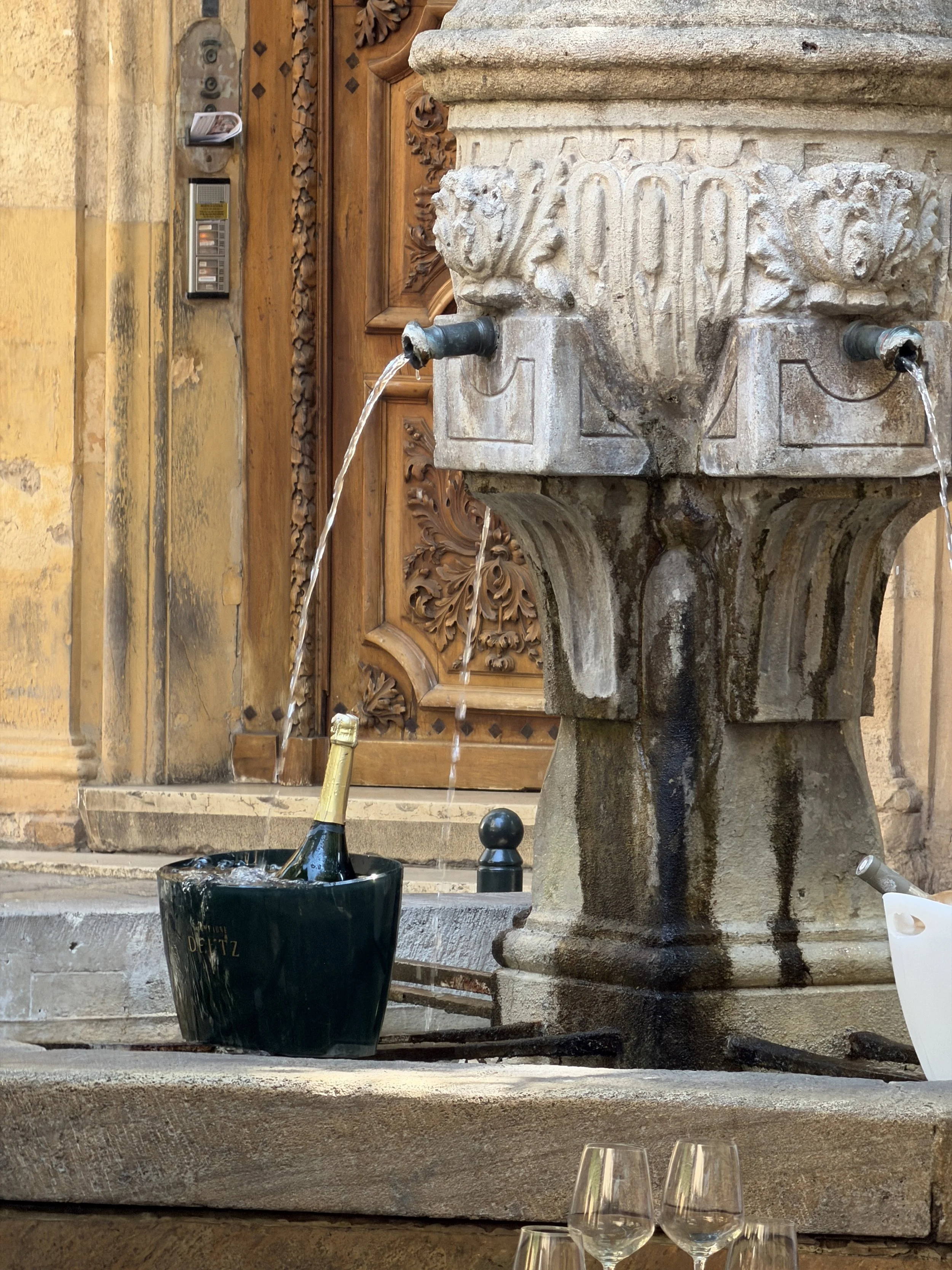 A stone fountain with two water spouts, one of which is pouring water into a black bucket with a champagne bottle inside. In front of the fountain, there are three empty wine glasses on the ground. The background features a wooden door and a decorative stone wall.