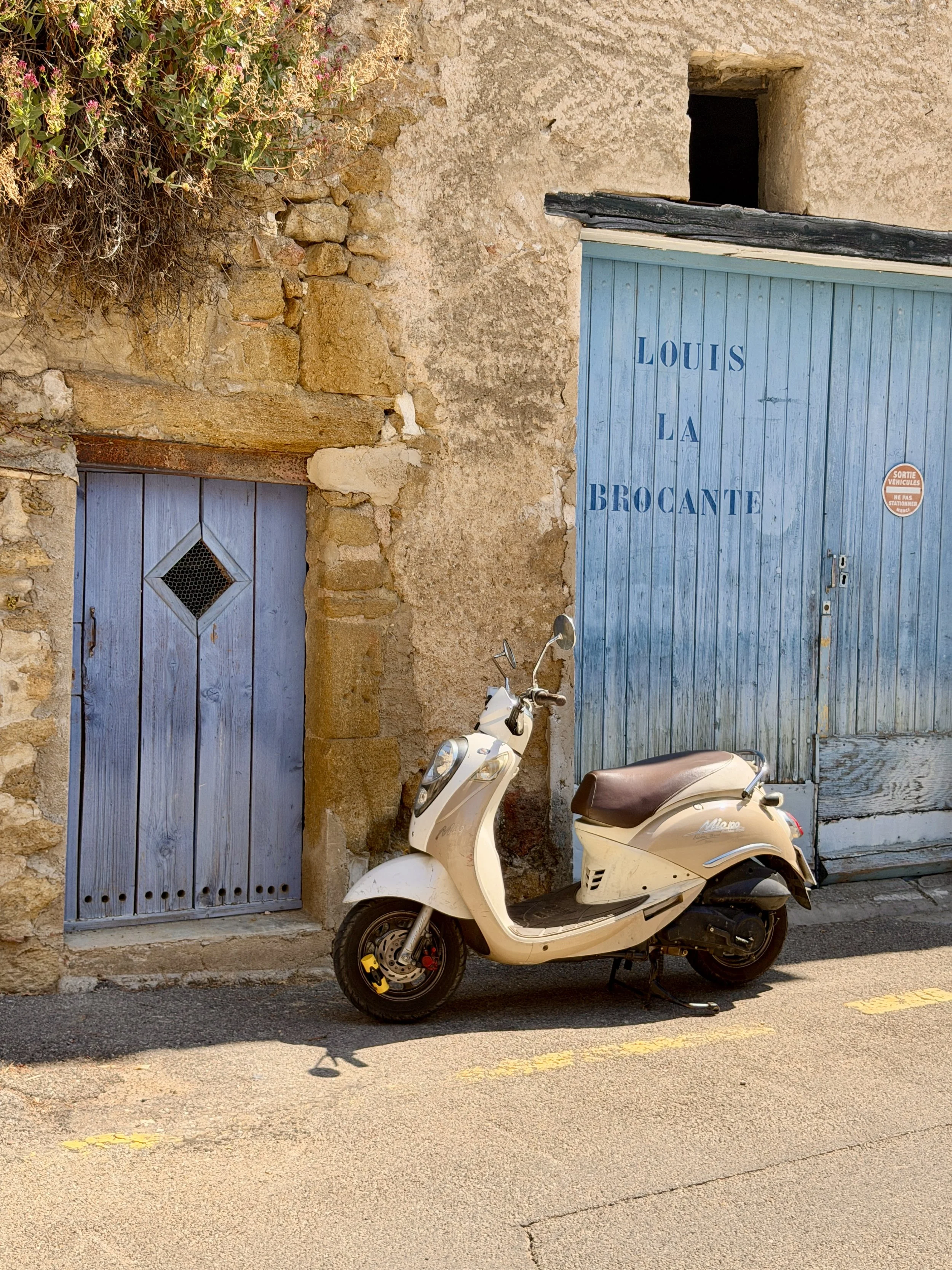 A beige scooter parked on the street in front of a rustic building with blue doors and stone walls, near a window with a small plant in the top left corner.