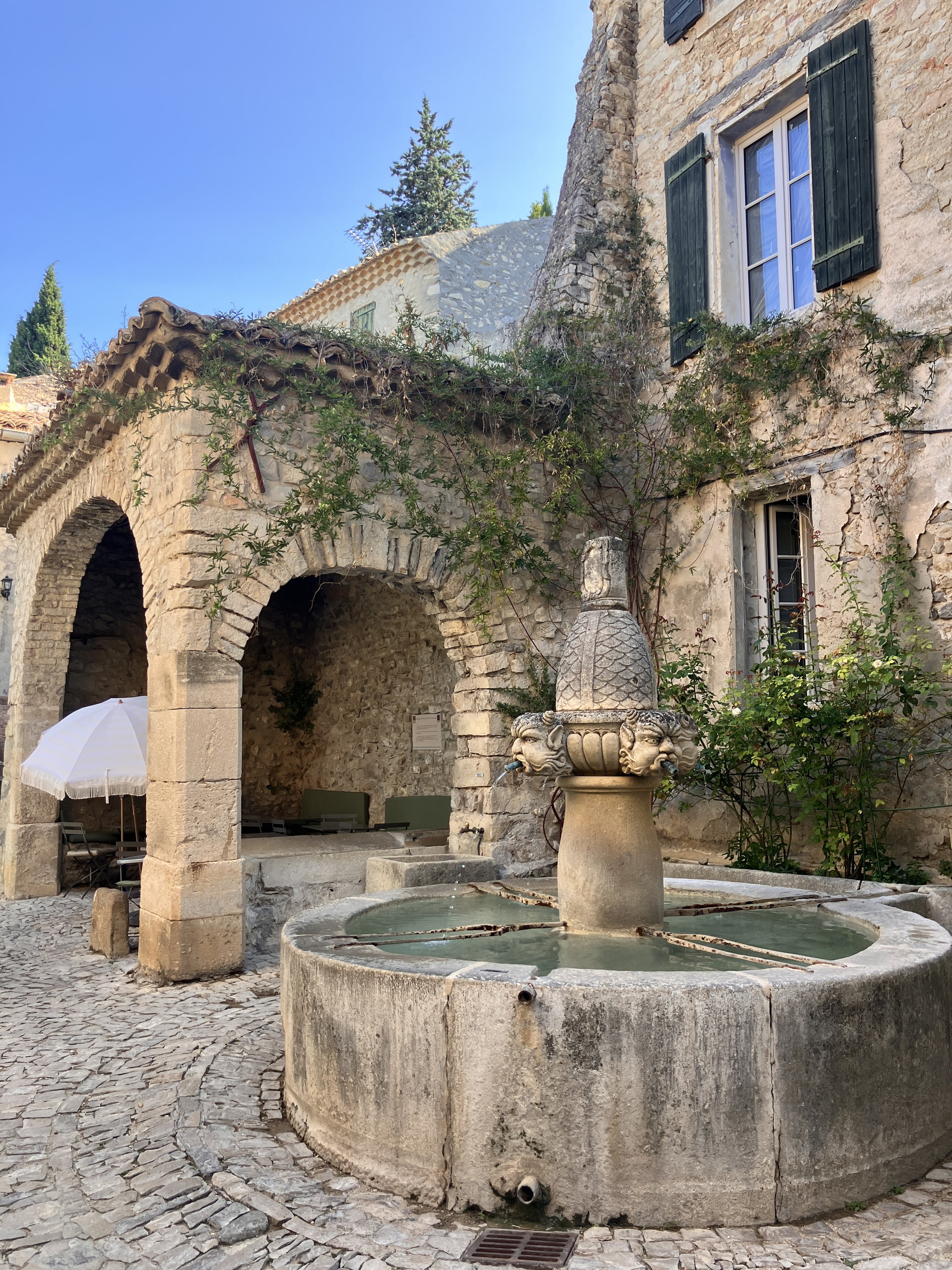 A stone courtyard features an ornate fountain with lion-head spouts, an archway with a brick and stone structure, and an old building with windows and green shutters. A white umbrella is partially visible in the bottom left corner.