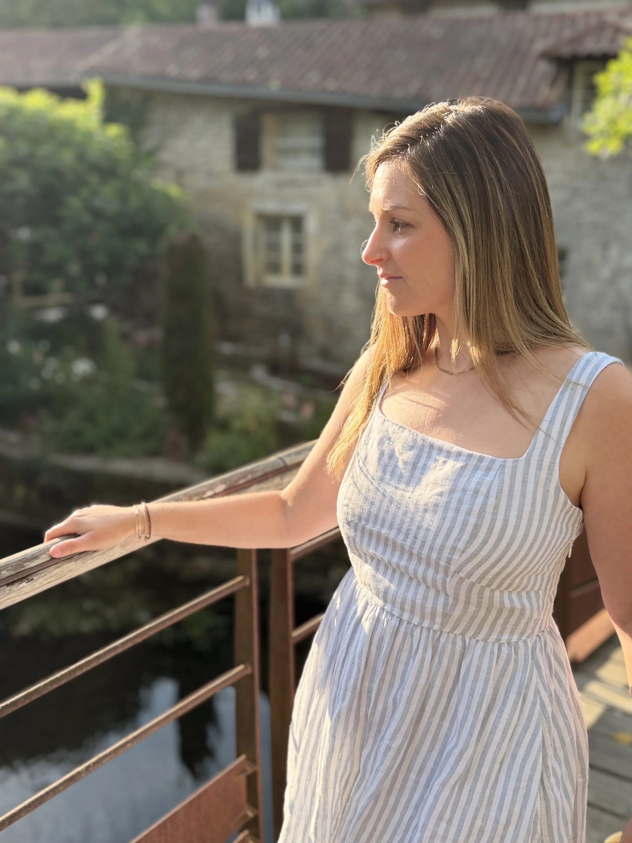 A young woman with brown hair and a nose piercing stands on a bridge, wearing a light blue and white striped sundress, looking to the right with a peaceful expression, in front of an old building with a tiled roof.