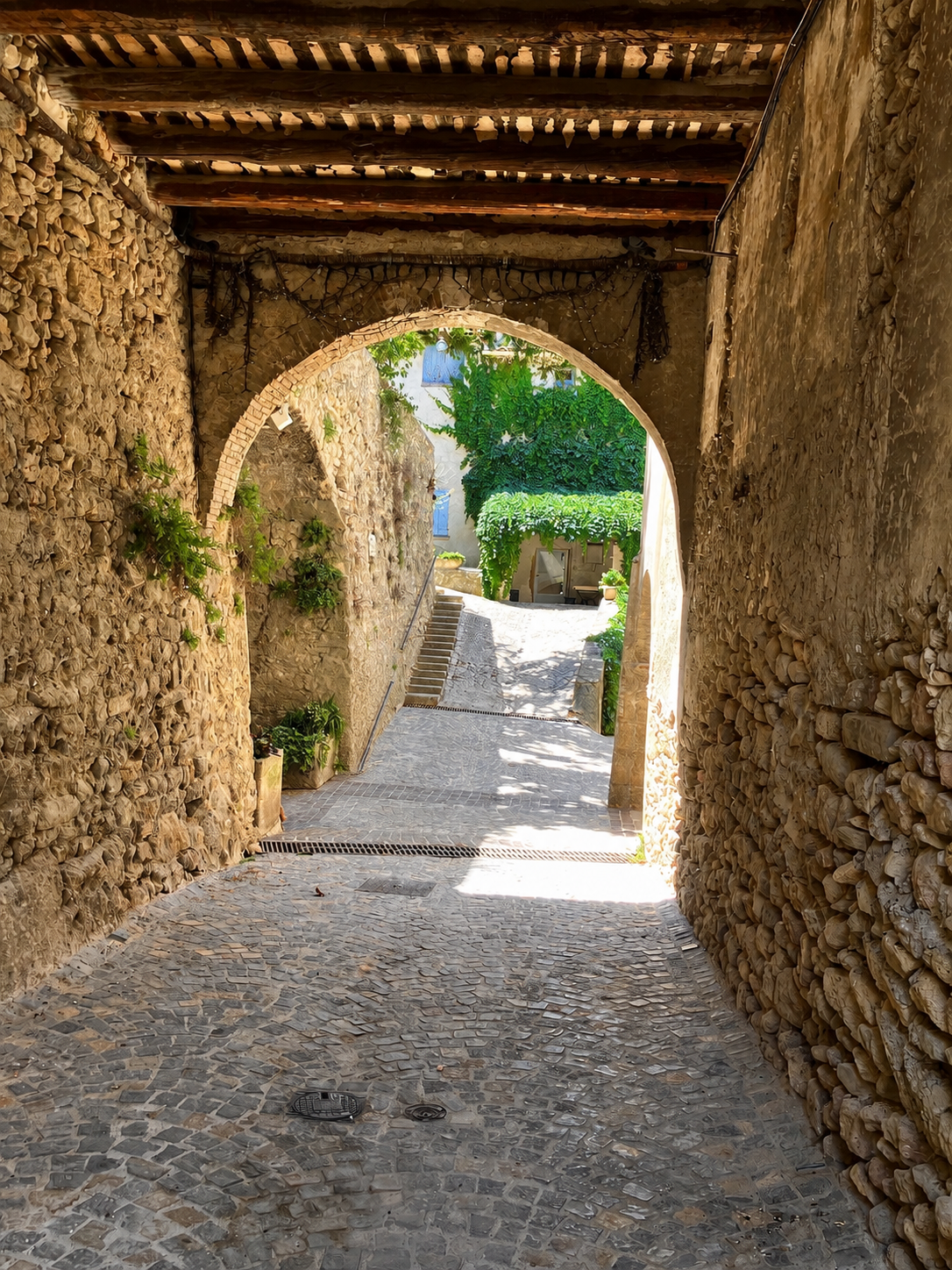Stone archway leading into a cobblestone alley with greenery and sunlight at the end.