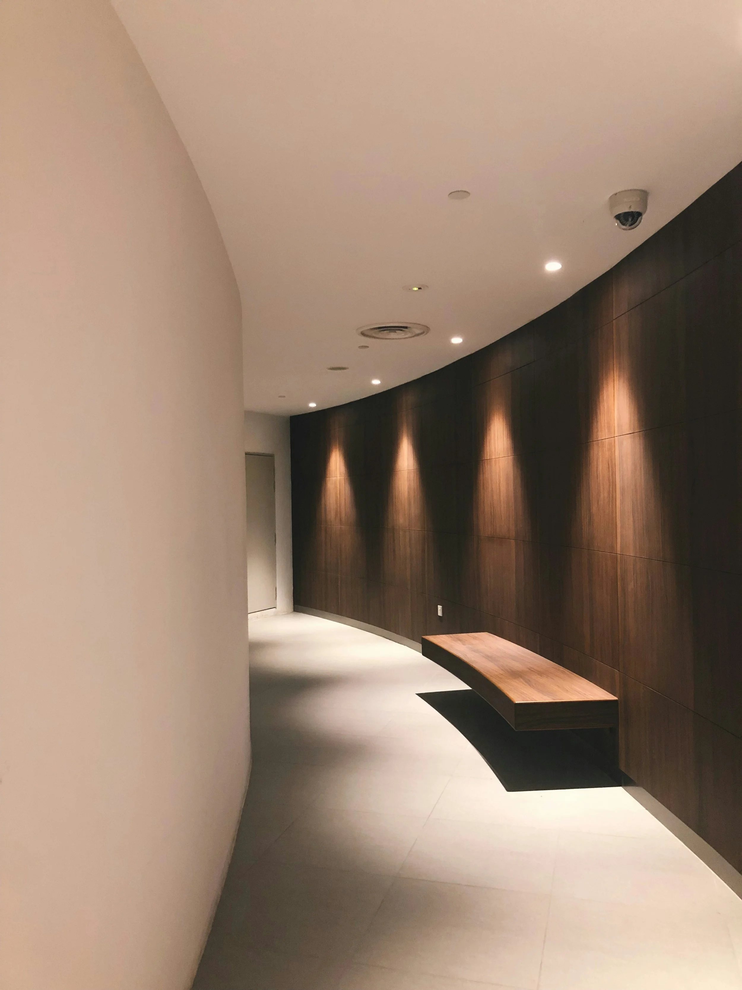 Interior hallway with white walls, dark wooden paneling on one side, a wall-mounted wooden bench, and ceiling lighting fixtures. A security camera is visible on the ceiling.
