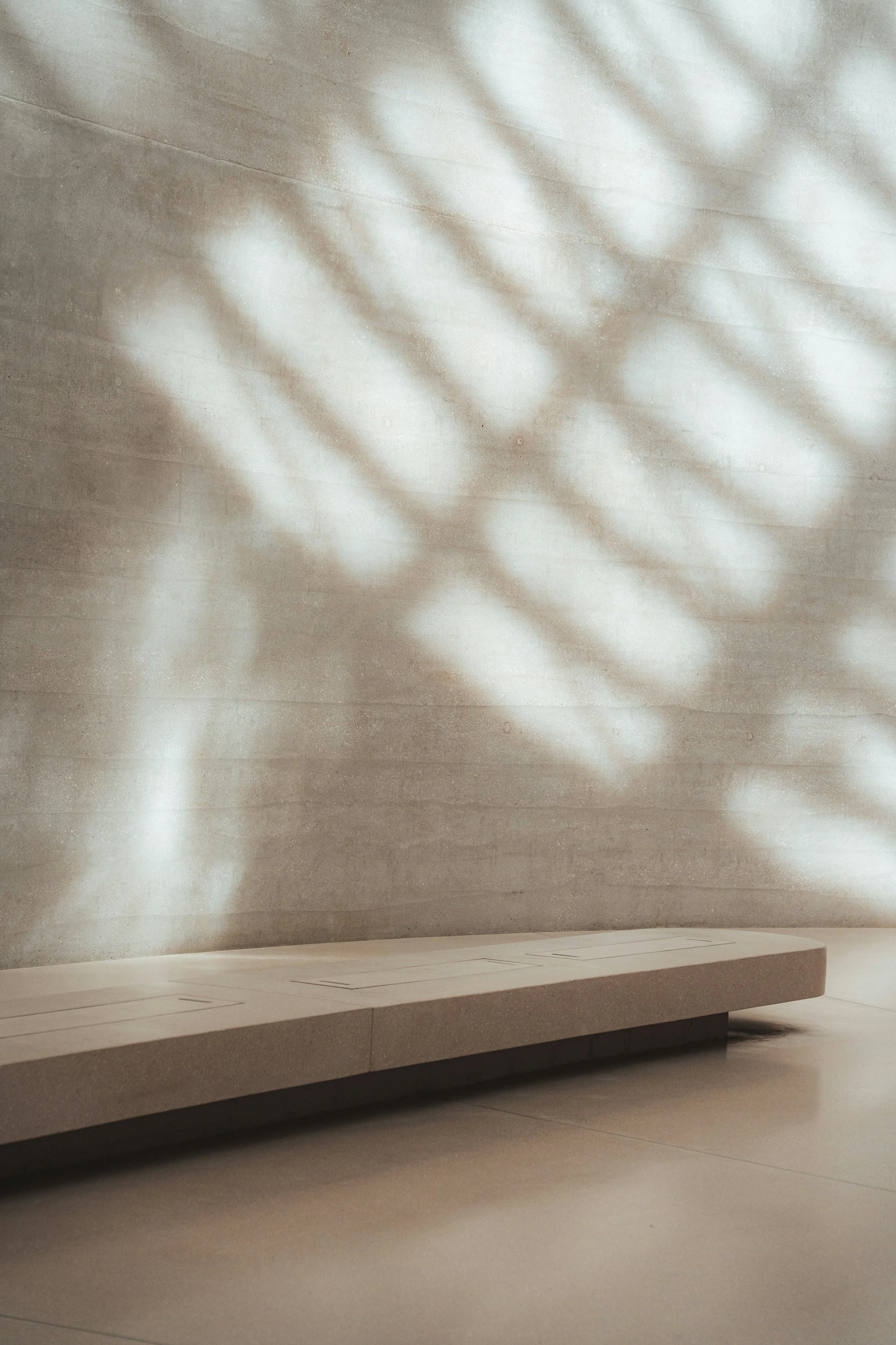 Minimalist modern interior with smooth, gray concrete wall, subtle lighting creating soft shadows, and a sleek white floating bench or platform.