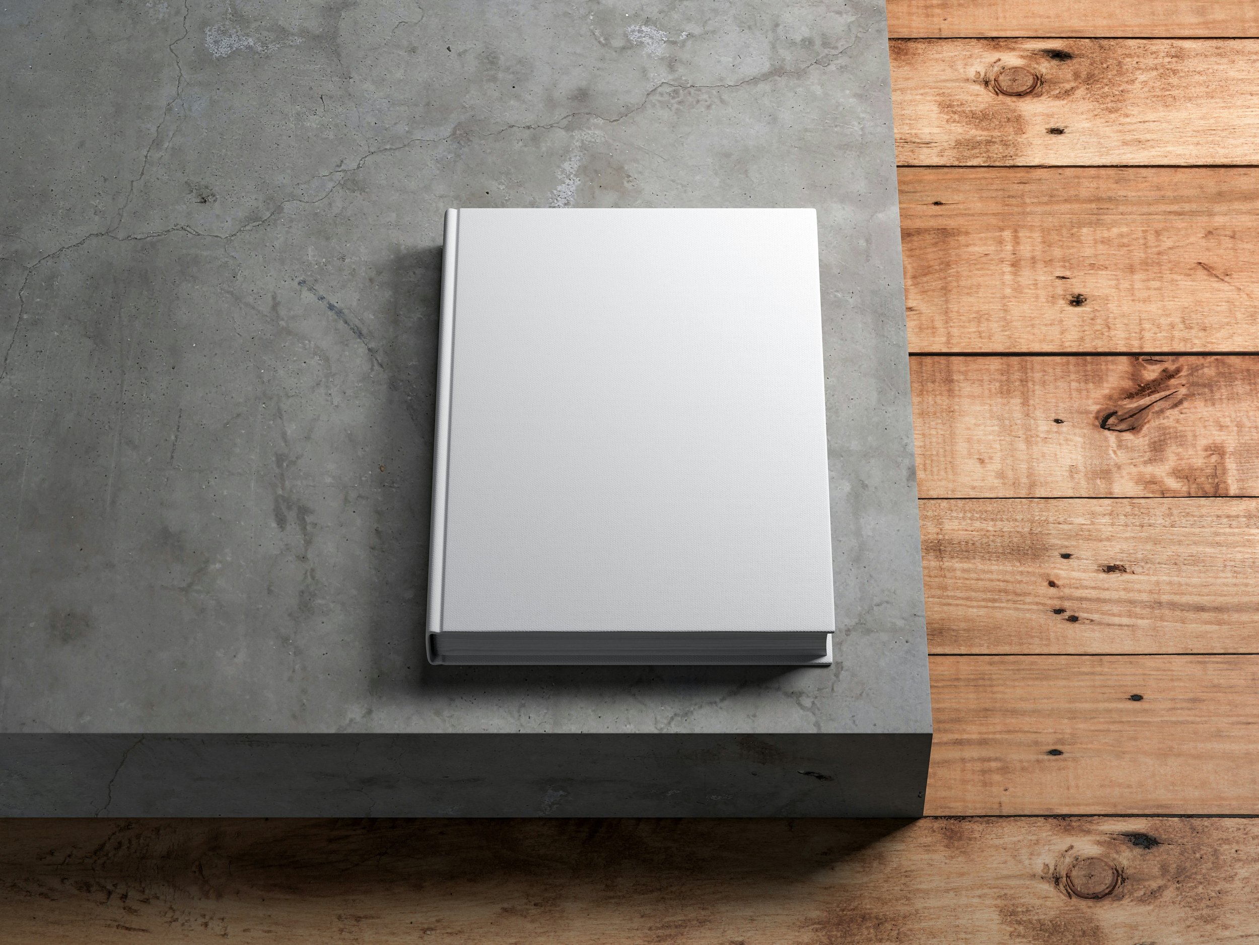 A white closed book resting on a surface that transitions from concrete to wood.